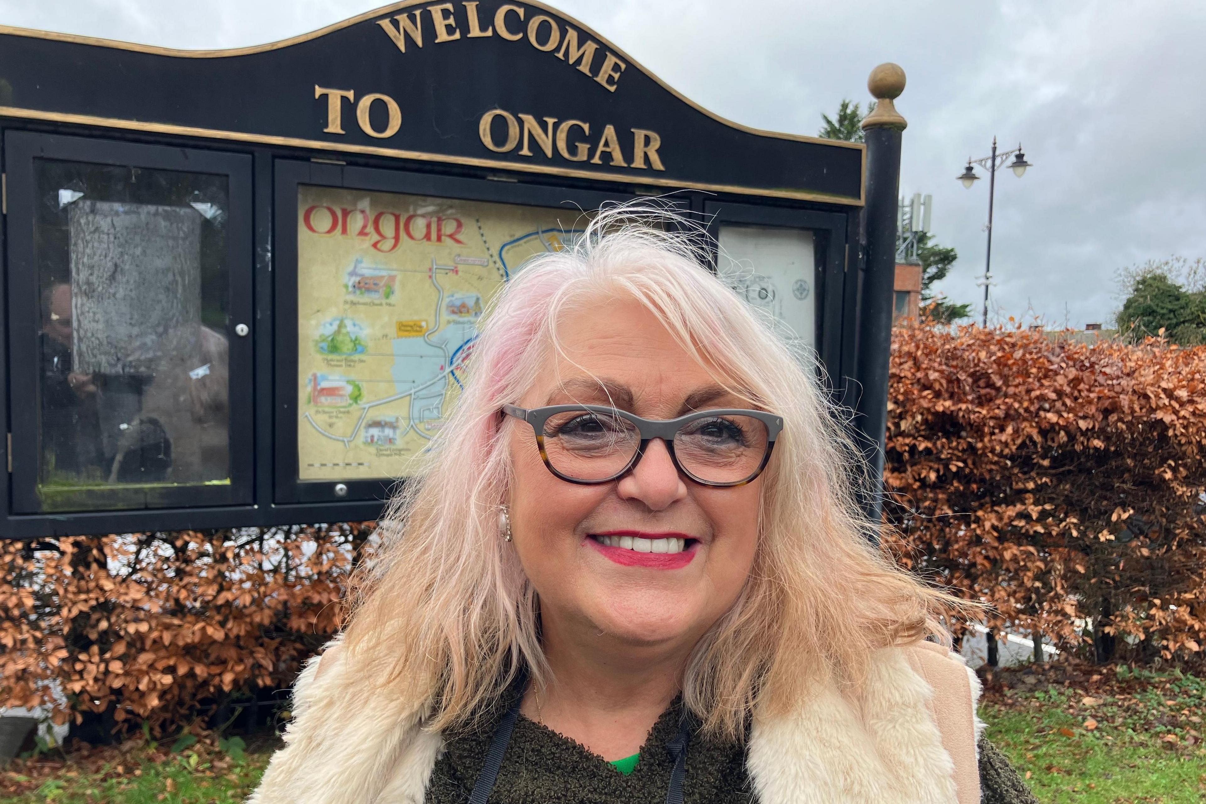 A head and shoulders picture of Barbara Hollingum - a resident of Ongar in front of the Welcome to Ongar town sign.