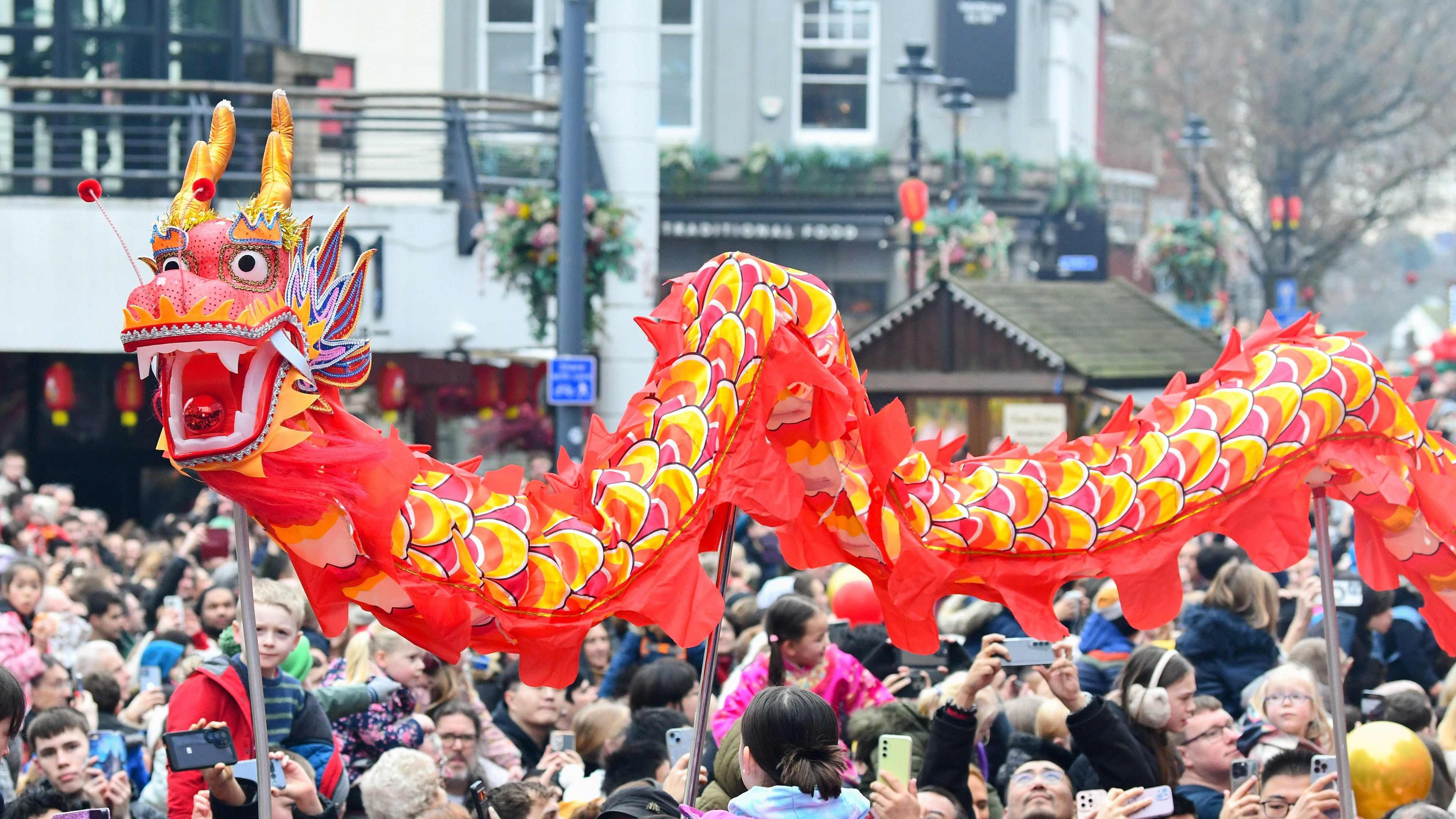 A red lunar dragon puppet with gold and orange detailing along the body. It is in a crowd of people who are holding up their phones and taking pictures.