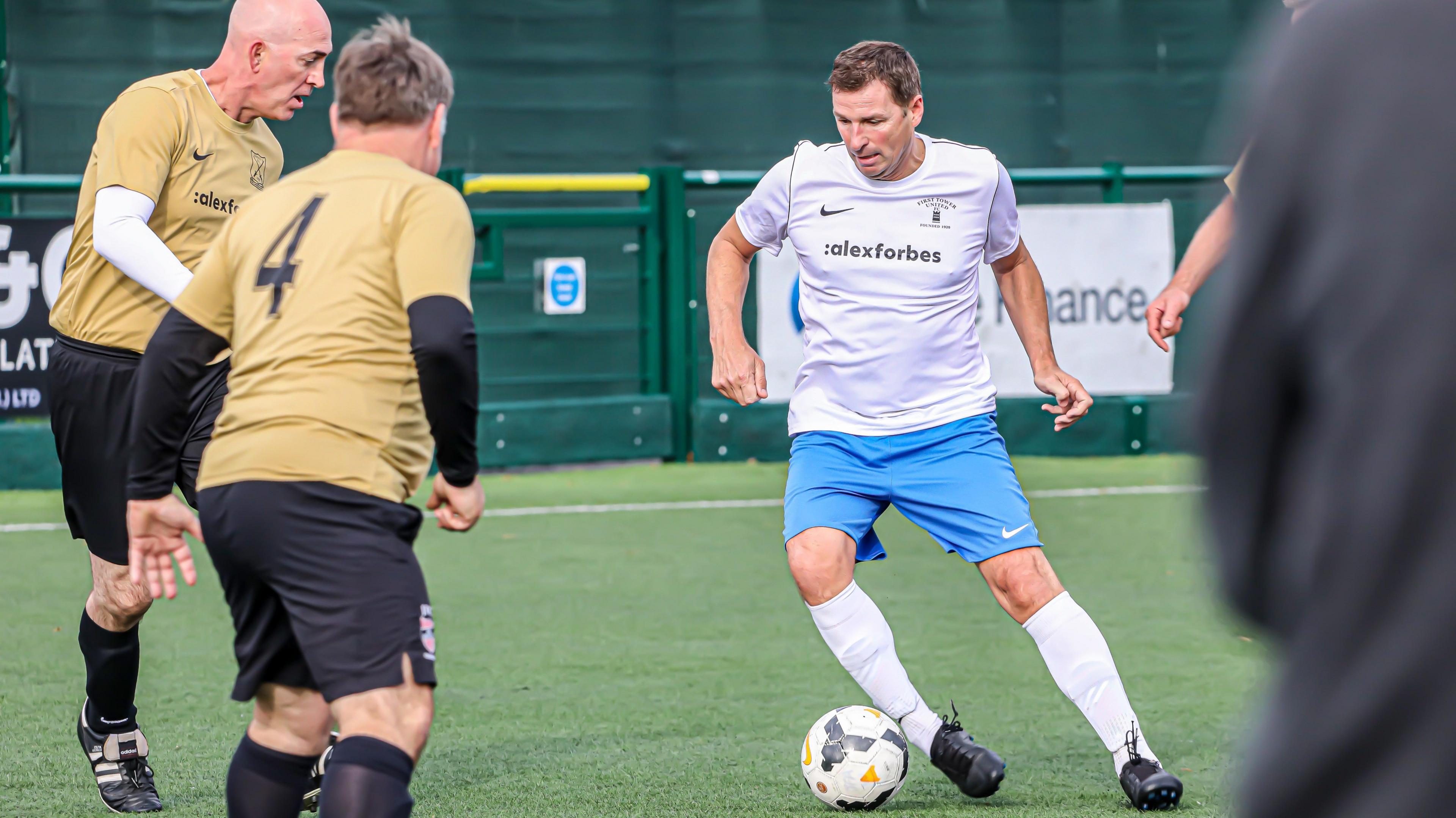 People playing walking football. A player in a white and blue kit is dribbling the ball at his opposition. two players are trying to block his path and are wearing black and gold kits. 