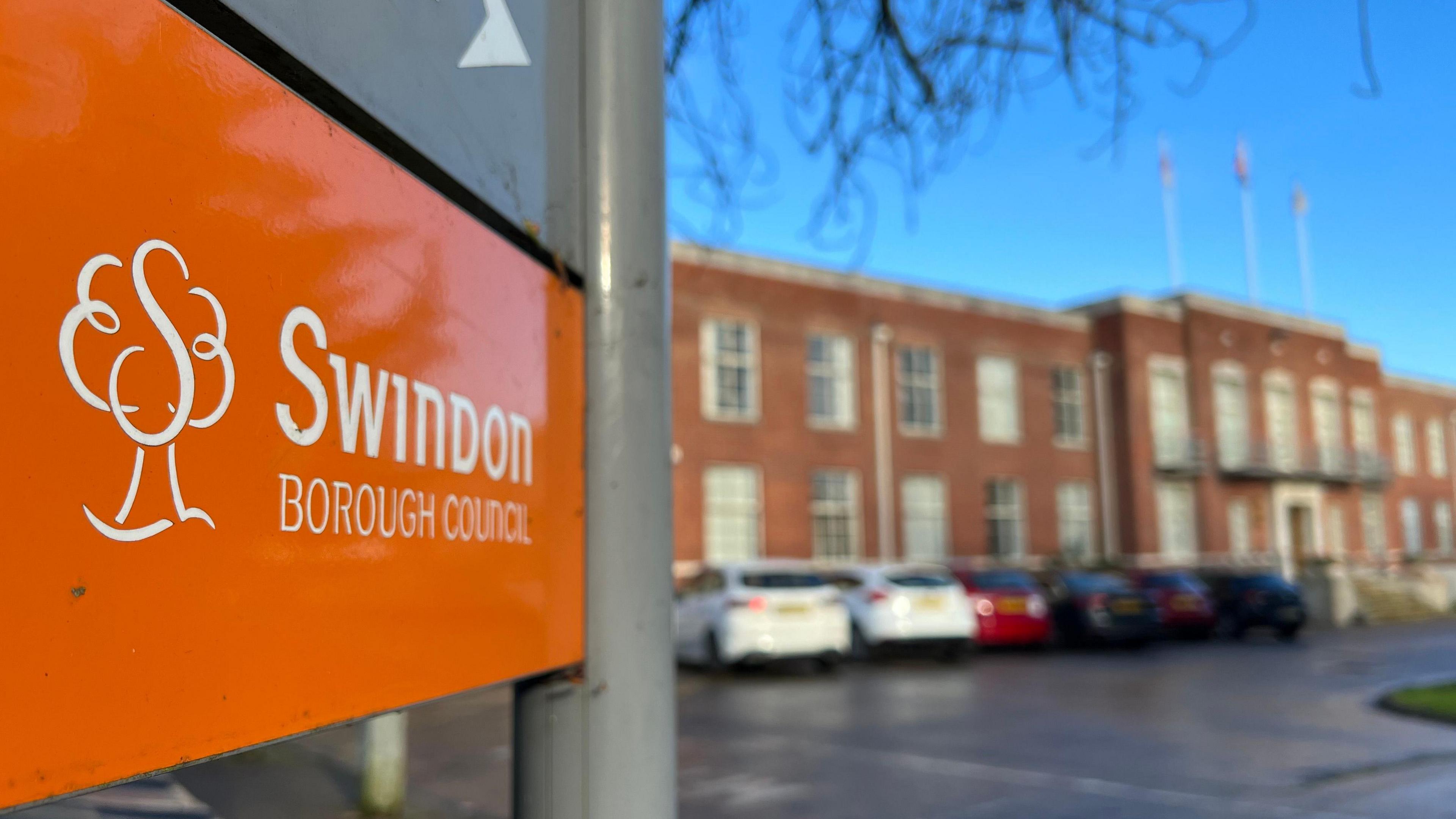 An orange Swindon Borough Council sign with a building behind it, with cars parked outside. The building in the background is out of focus.