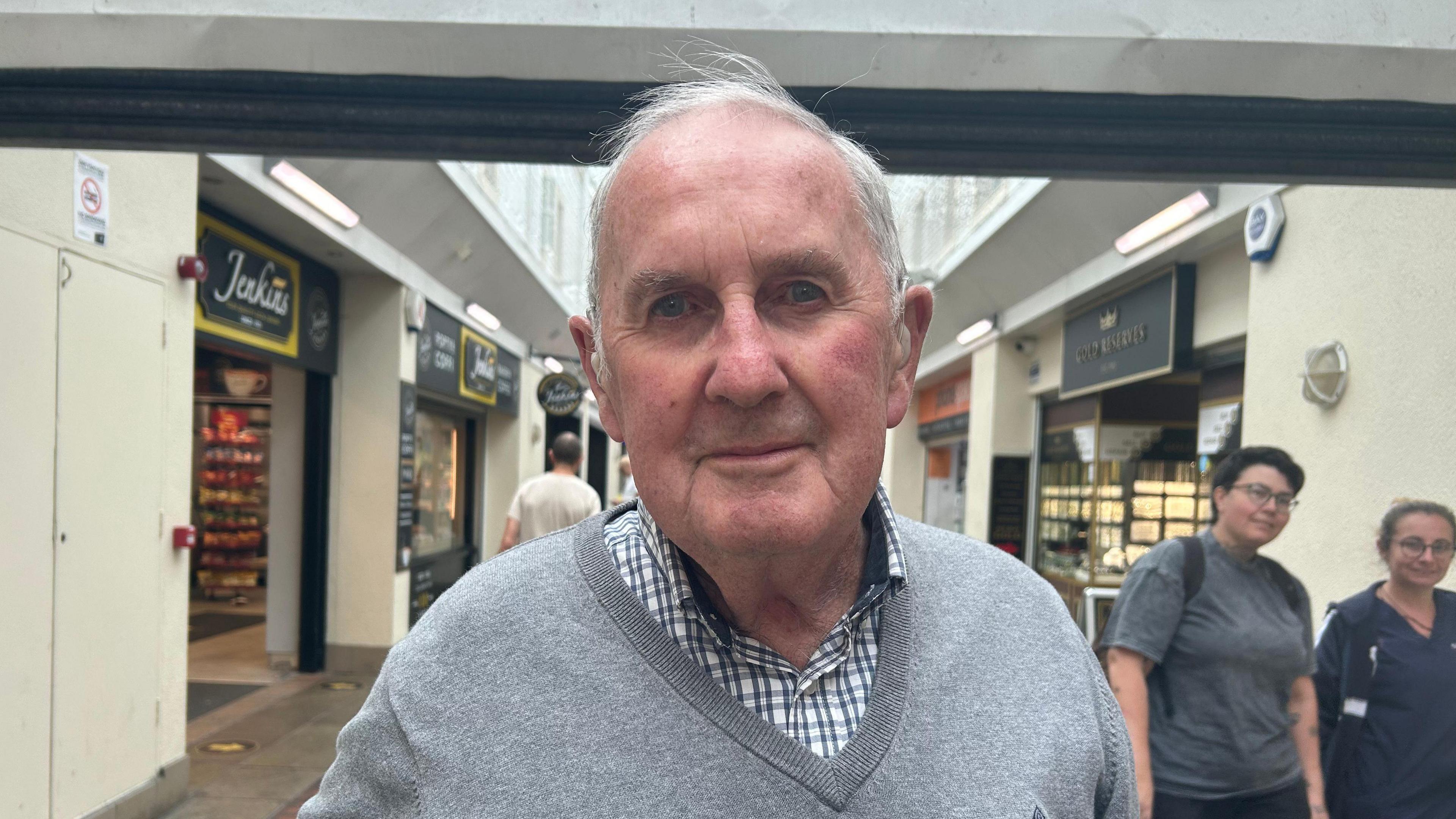 Alan smiling looking at the camera whilst standing in Llanelli town centre. He is wearing a grey v-neck jumper and a blue and white tartan shirt. He has short grey hair.