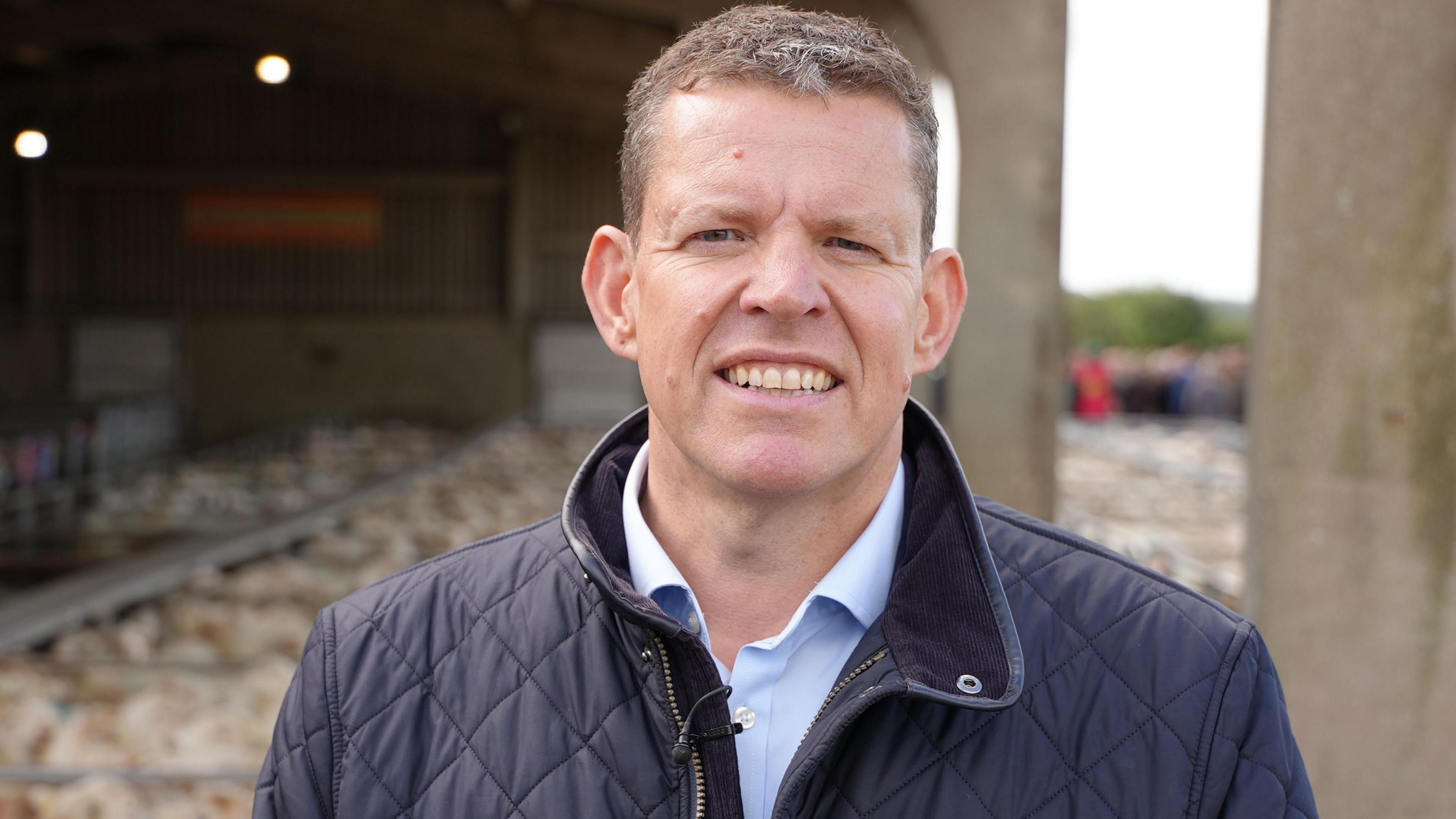 Rhun ap Iorwerth smiling, wearing a blue coat with a light blue short underneath with sheep in pens behind him at Gaerwen farmers market, Anglesey