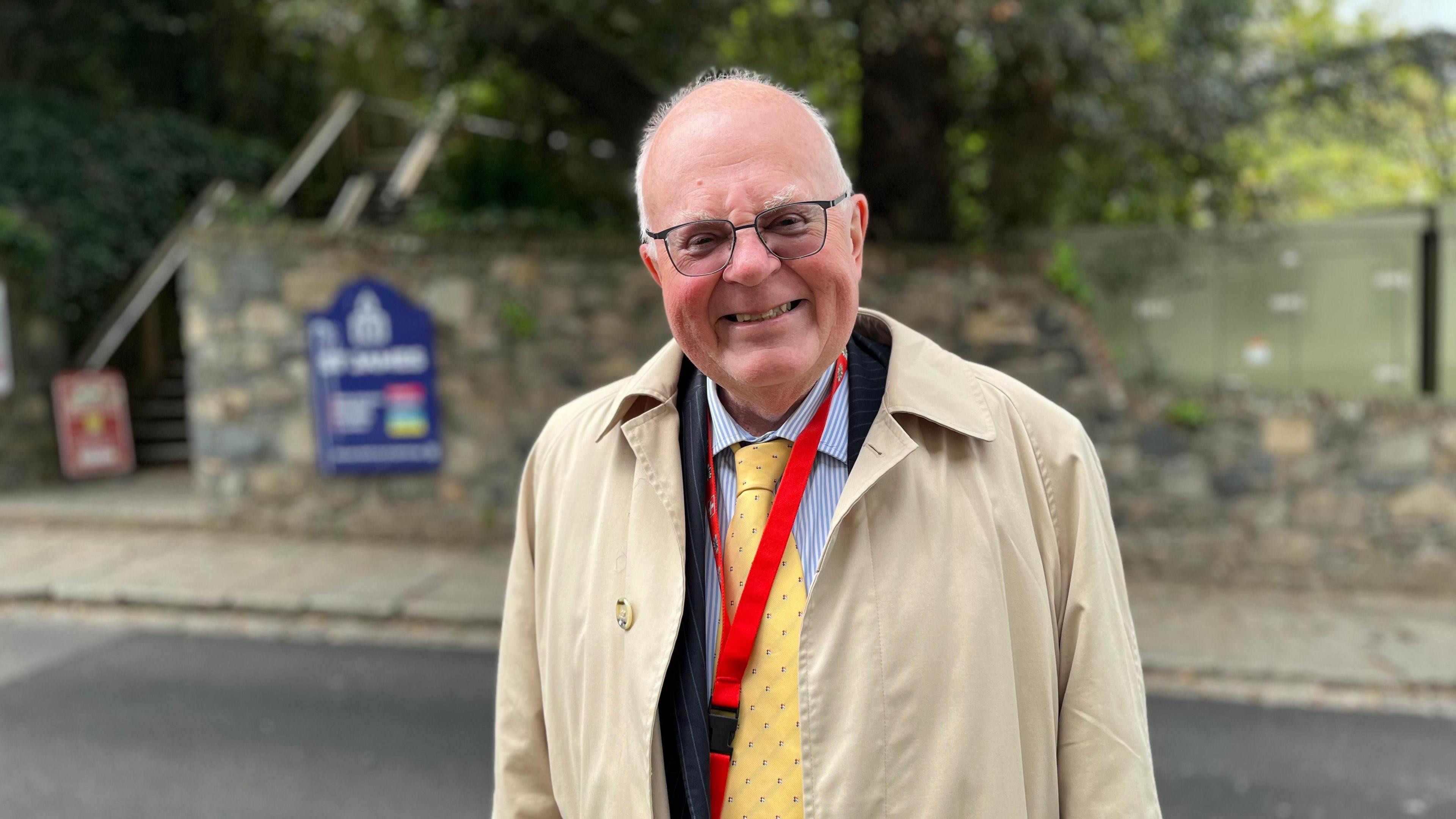 A partially bald man wearing a long, light brown coat, a blue suit and a yellow tie stands next to a road with an urban park behind him.