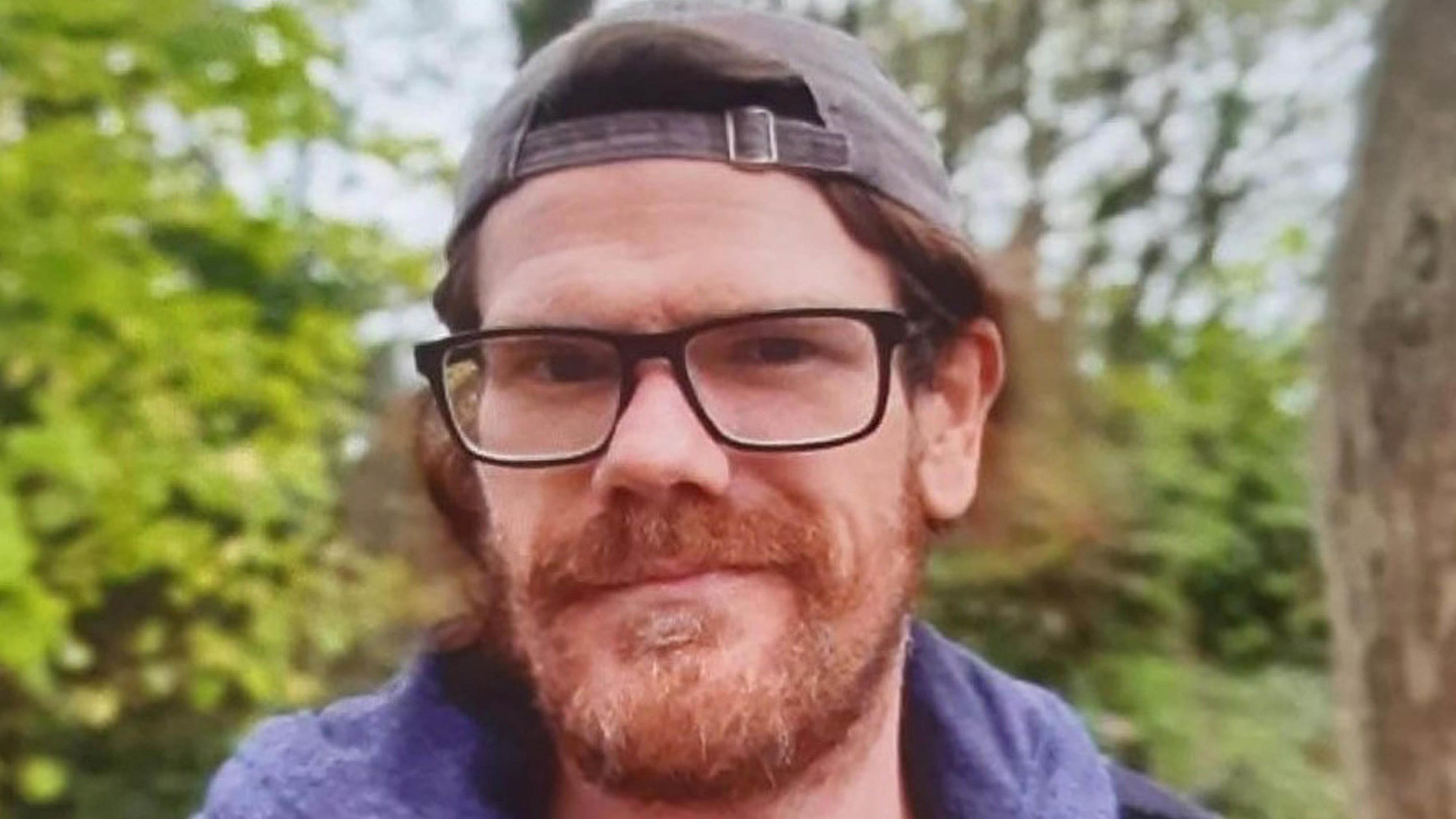 Shane Lynch is smiling at the camera as he stands outside with foliage behind him. He has a ginger brown beard and collar-length hair. He is wearing a baseball cap backwards and a pair of brown rimmed glasses.