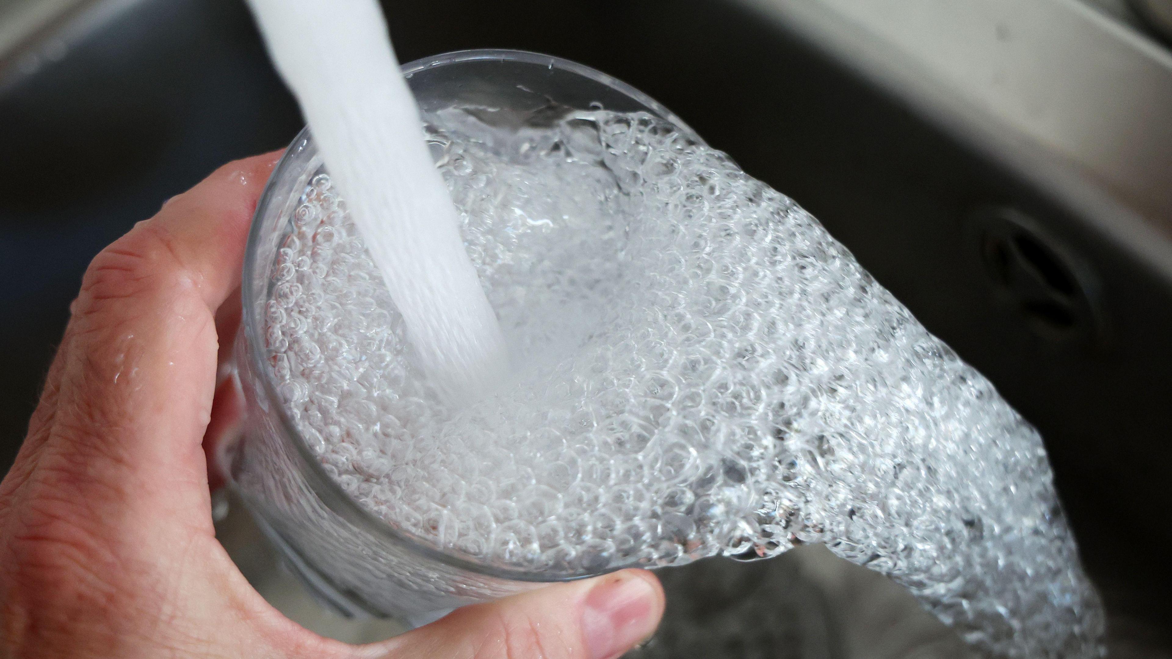 A hand holding a glass under a running tap.  The water is full of bubbles and is overflowing into a chrome sink.