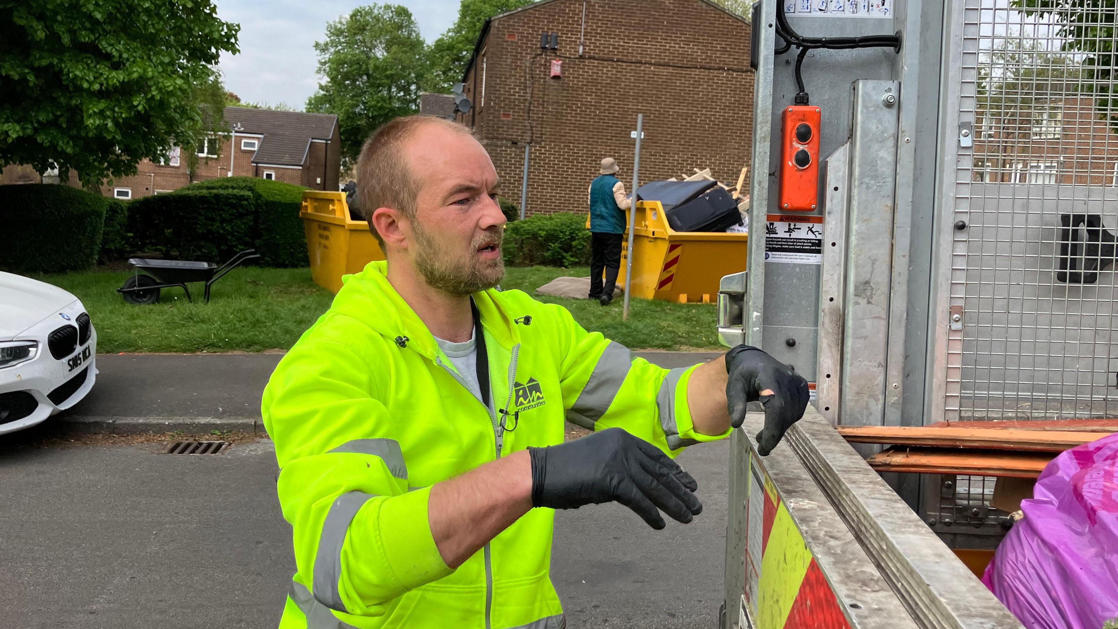 A man in a yellow high visibility vest and wearing black gloves is stood next to a van full of rubbish