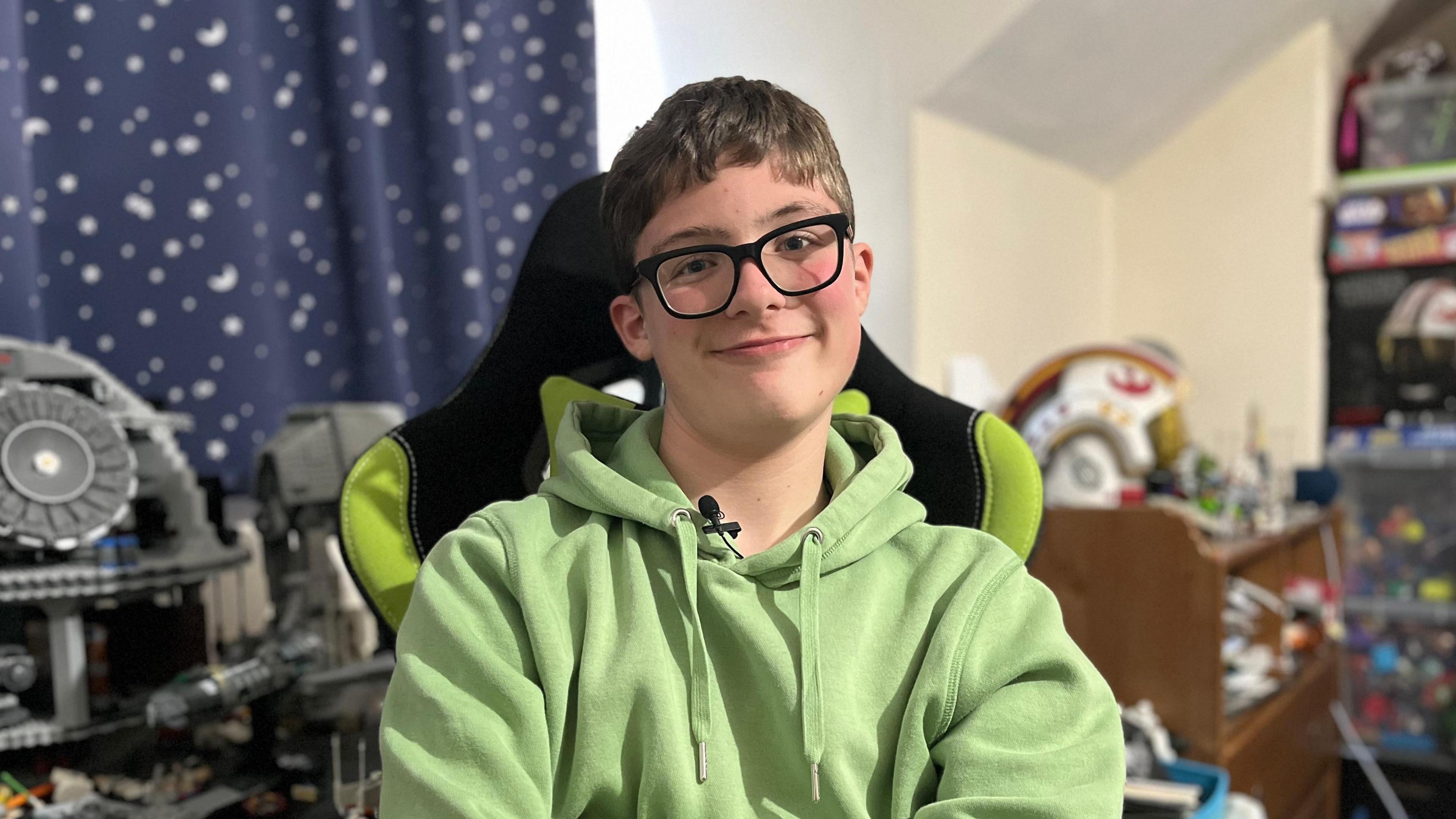 Liam is staring directly at the camera and wearing a light green hoodie. He is sat in a desk chair that is black and green - and although the background is blurred, Lego and Star Wars toys can be seen. He is wearing black rimmed glasses and he has short brown hair. 