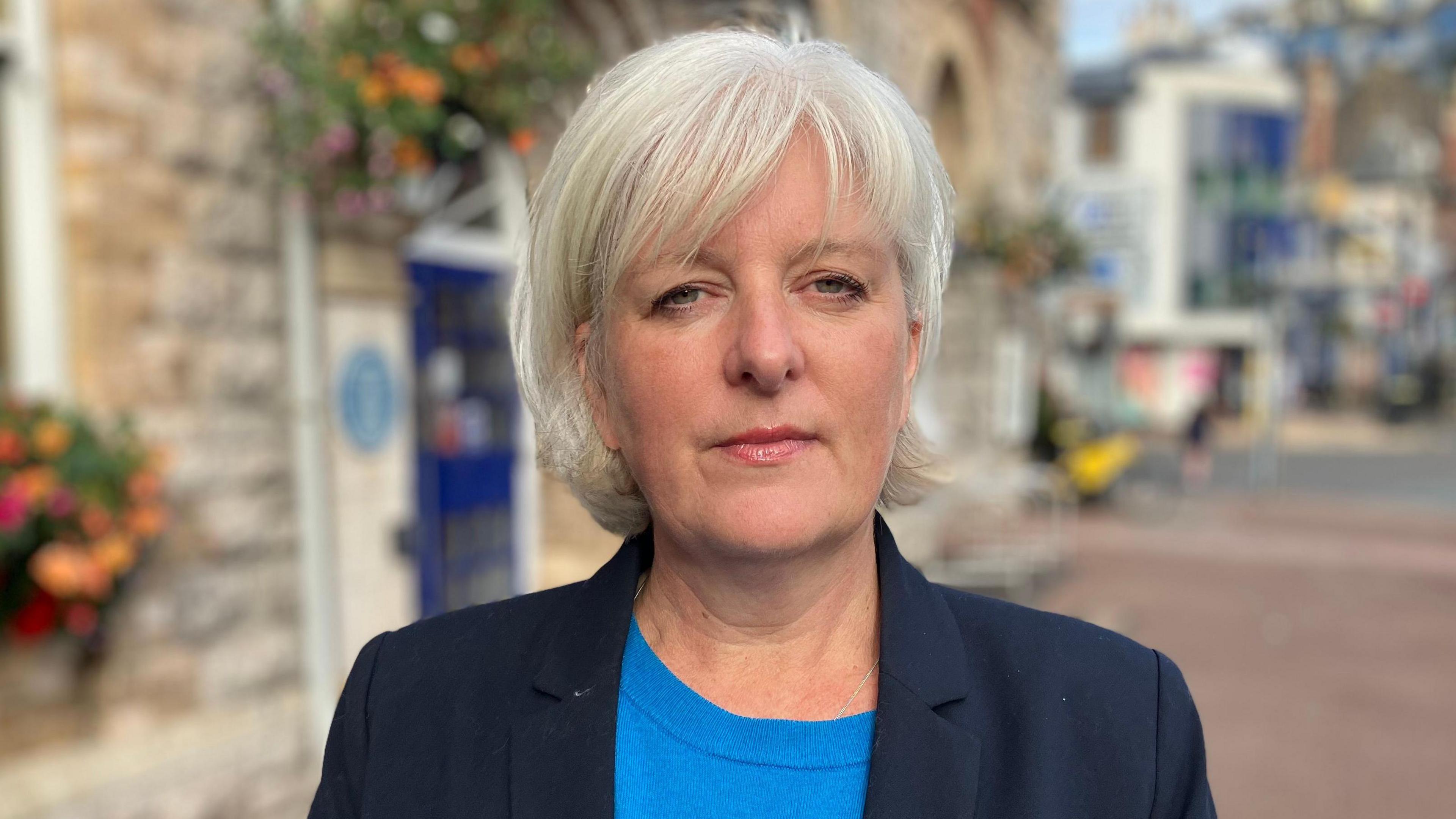 South Devon MP Caroline Voaden wearing a navy blue blazer and light blue top. She is standing in front of a building decorated with planters full of flowers on a street in a town centre.