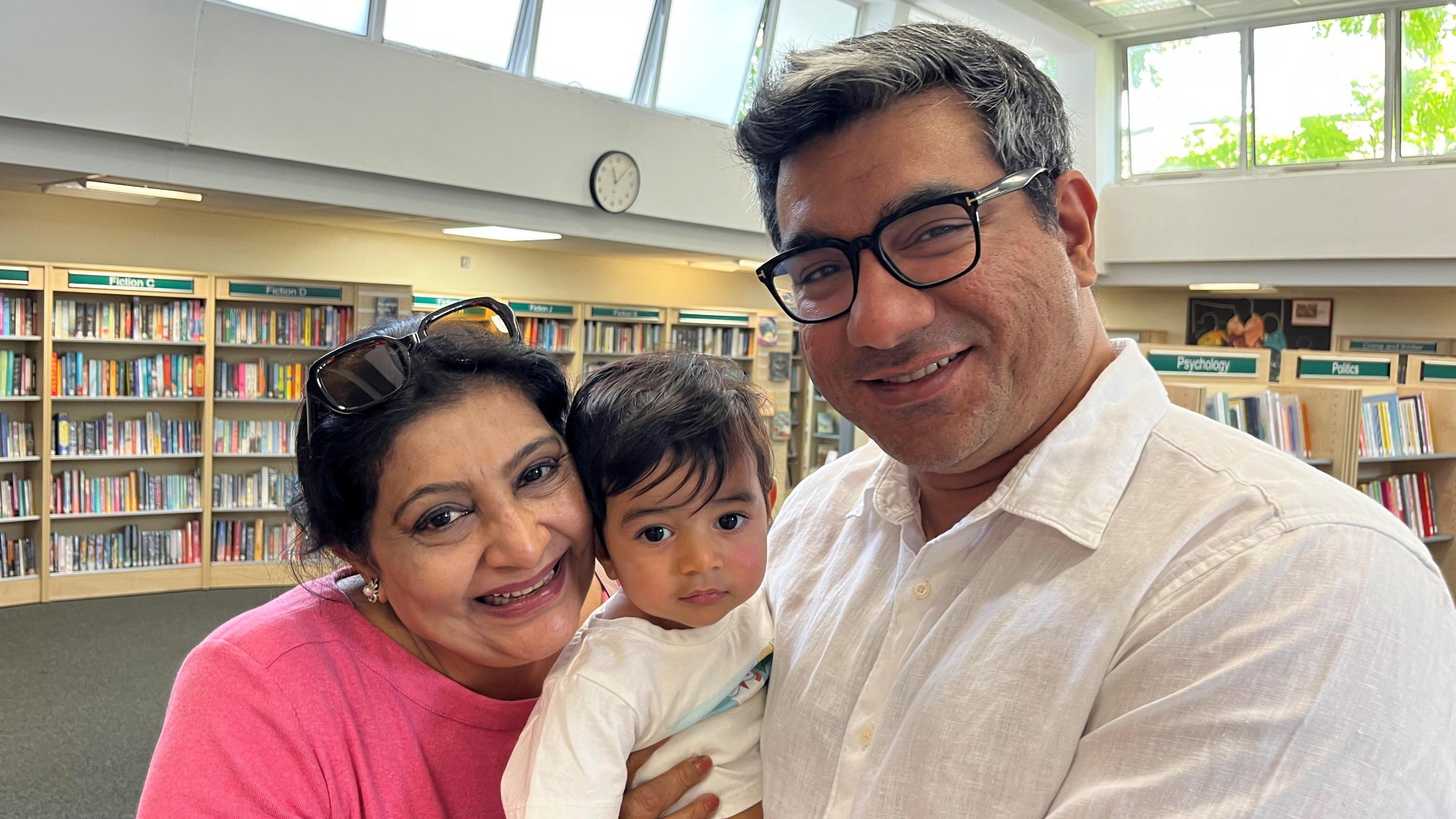 Aasim Hussain on the right holding his toddler son Armaan in the middle of the shot, and his son’s grandmother Fouzia is on the left. They are in Brompton Library in Kensington