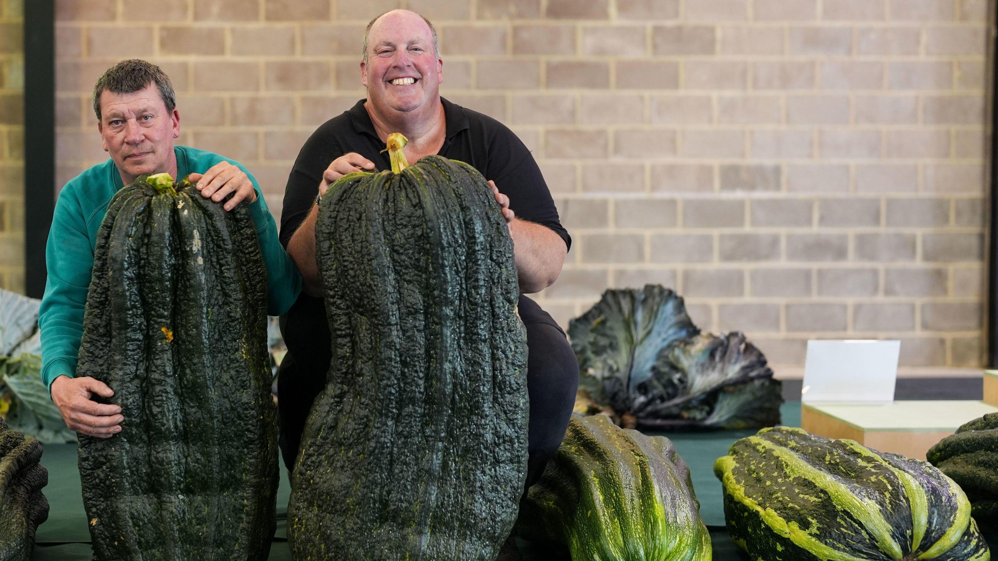 Two men, one in a green jumper and the other in a black t-shirt, hold up oversized marrows.