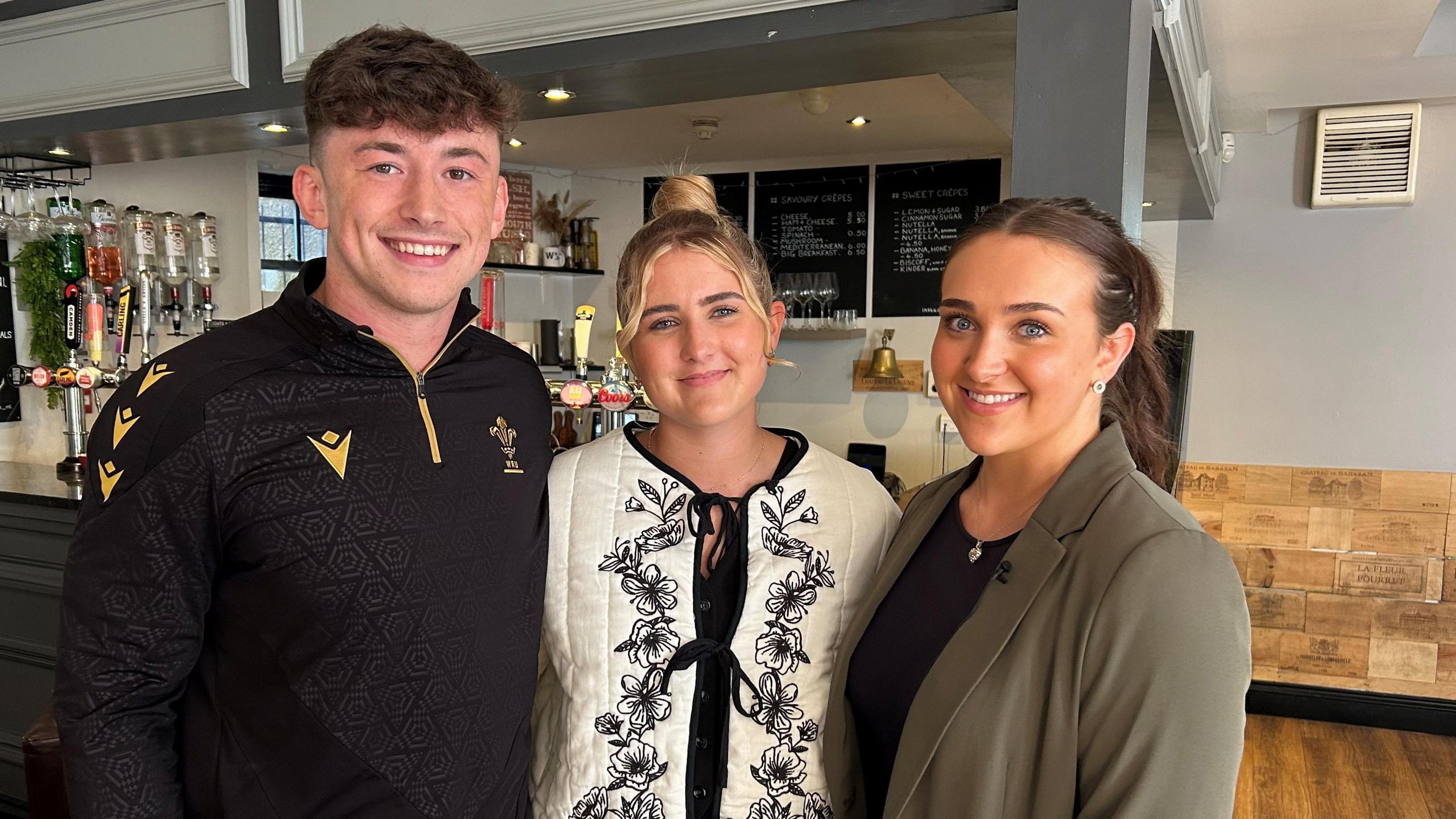 An image of me, Finley and Bronnie stood in front of a bar at our local pub. We are all smiling looking at the camera. Finley, on the left, is wearing a black quarter zip sports top. He has short brown hair. I am in the middle, I am wearing a cream and black padded patterned jacket. My hair is in a top bun. Bronnie, on the right, is wearing a green blazer and black top. Her brown hair is in a high ponytail.