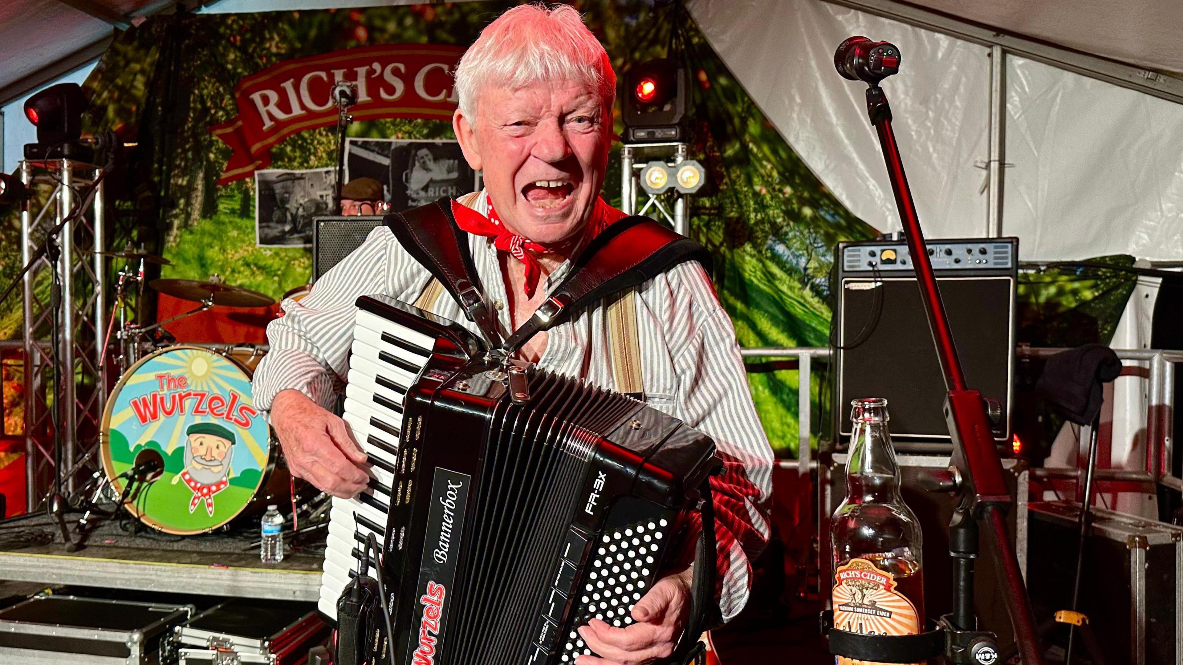 Accordian player from the Wurzels wearing a red scarf on his neck singing at the camera with the drummer in the background