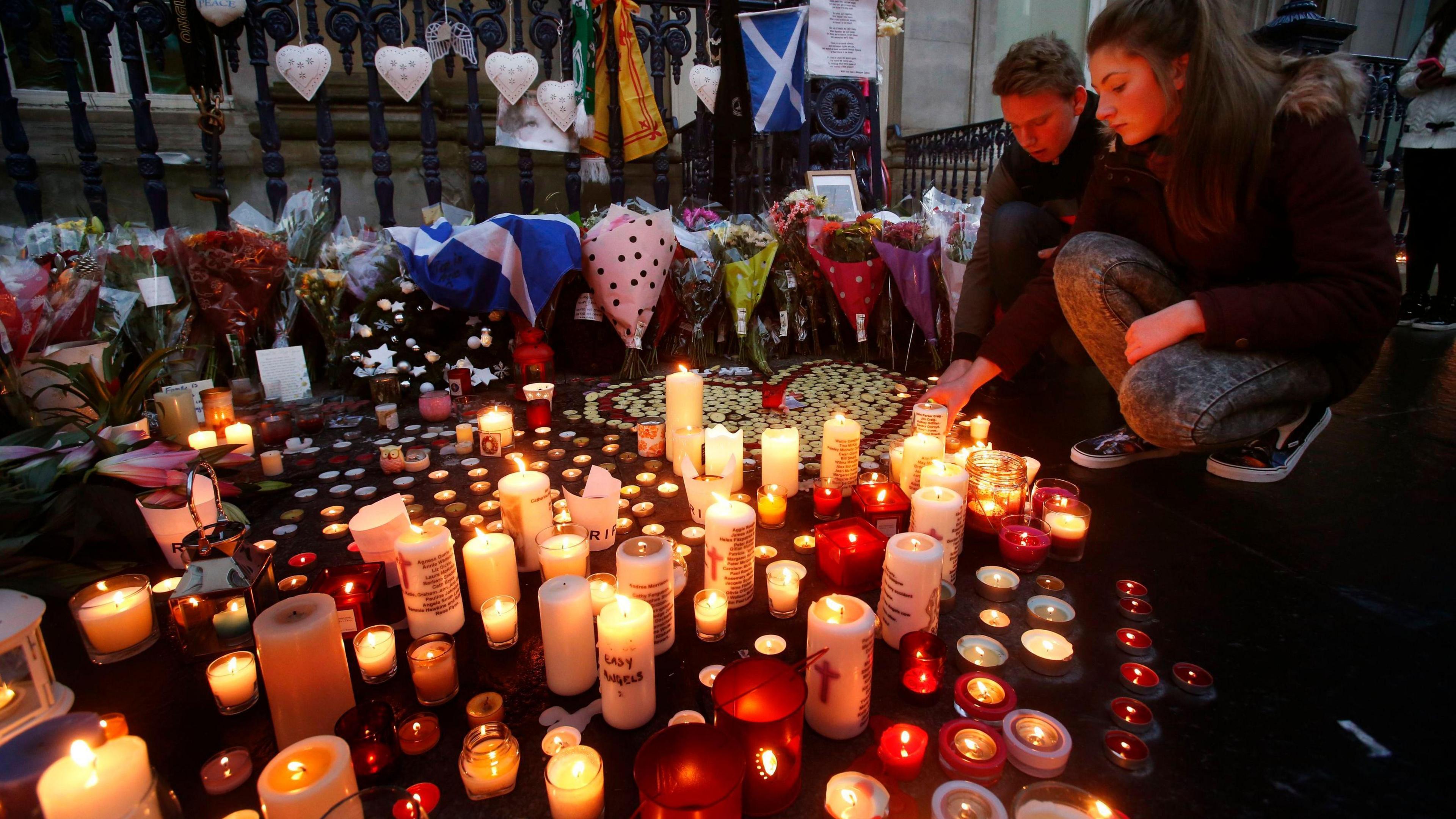 A young man and woman crouch down to light candles on a pavement in Glasgow city centre in tributes to the bin lorry crash victims. Flowers and messages of condolences are propped up against a fence, near the candles.