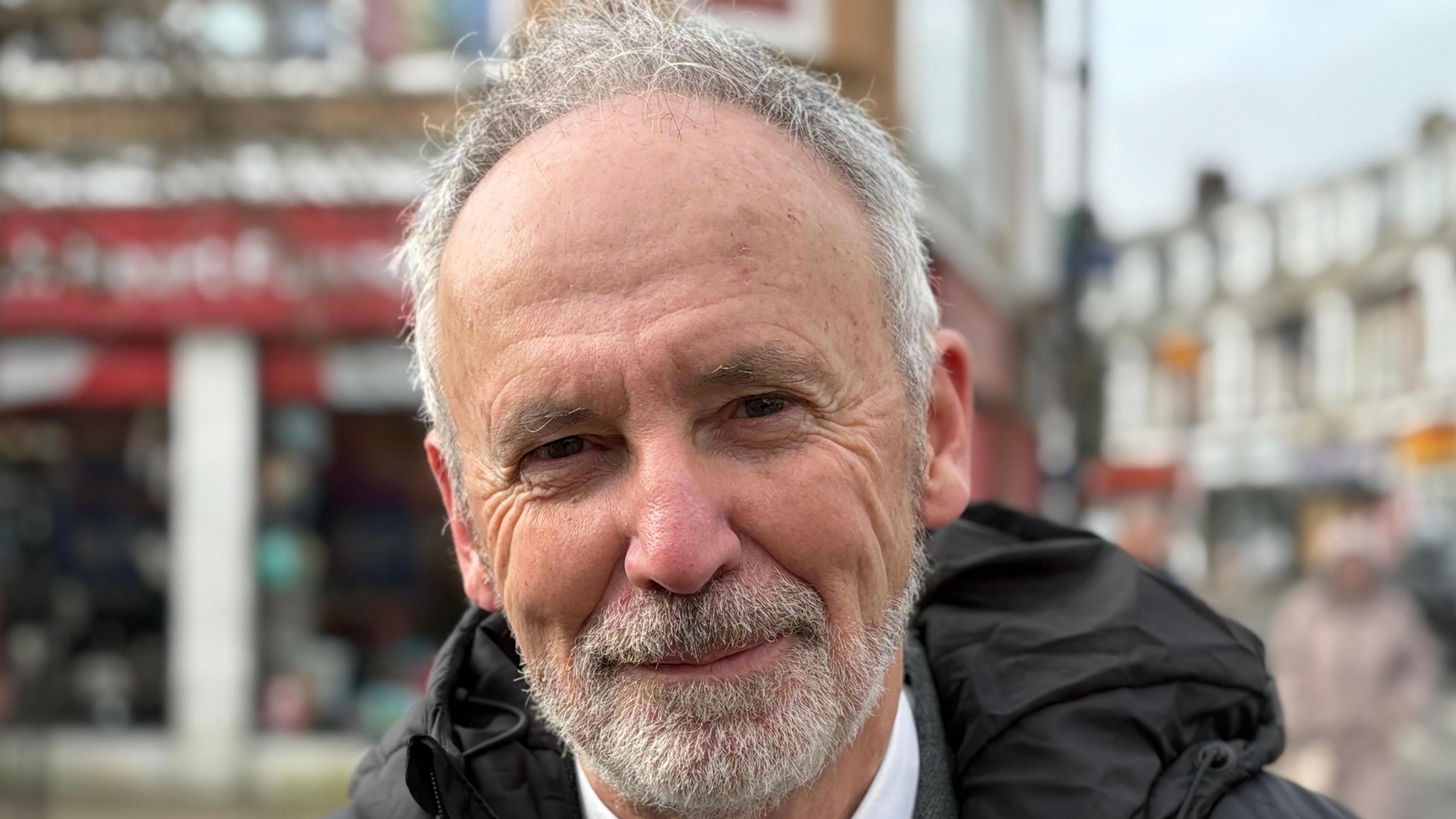 A head shot of John Cosgrove. He has grey hair and a grey beard and is smiling directly at the camera. There is a street scene behind him.