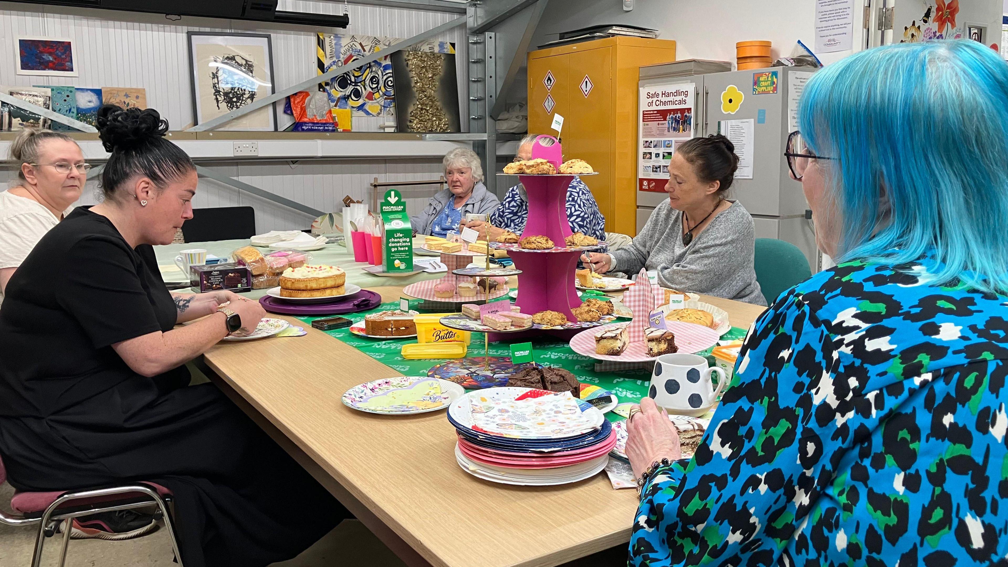 A group of six women sit around an oblong table. In the centre is a pink, tiered cake stand covered in scones, with other cakes on plates around the table.  In the background is framed artwork on the walls, a tall yellow cupboard and a tall grey cupboard. 