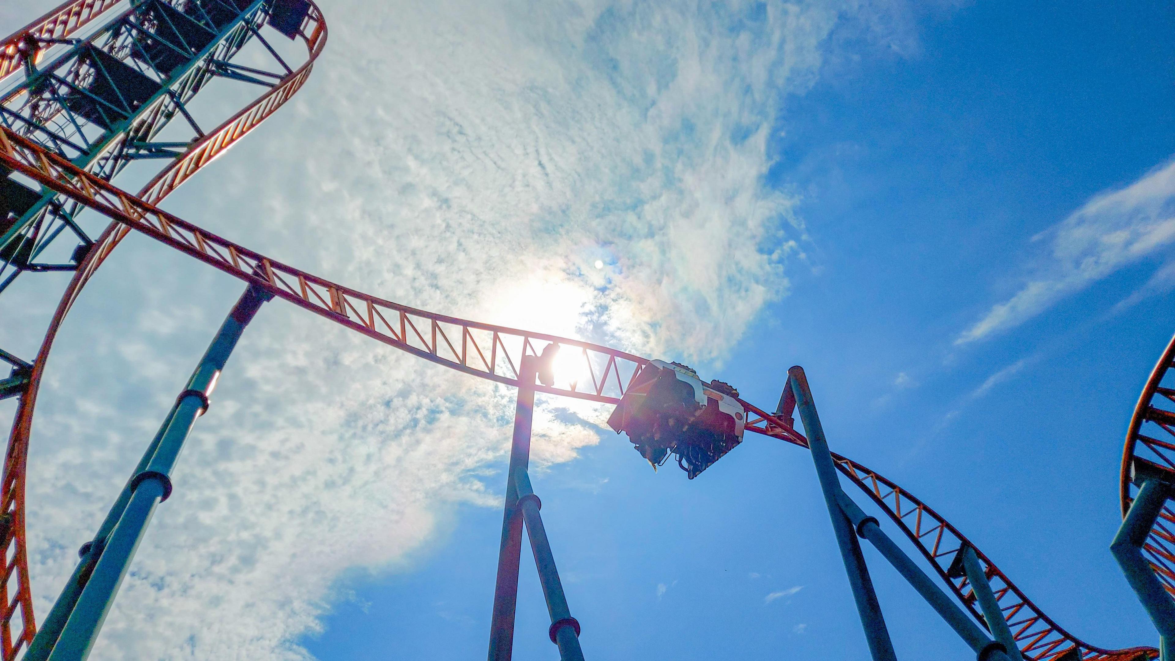 A rollercoaster pictured from below. You can see a cart upside down making its way down a rotating track. The rollercoaster has an orange track with blue support beams. Behind it is a blue sky with the sun shining through a cloud. 