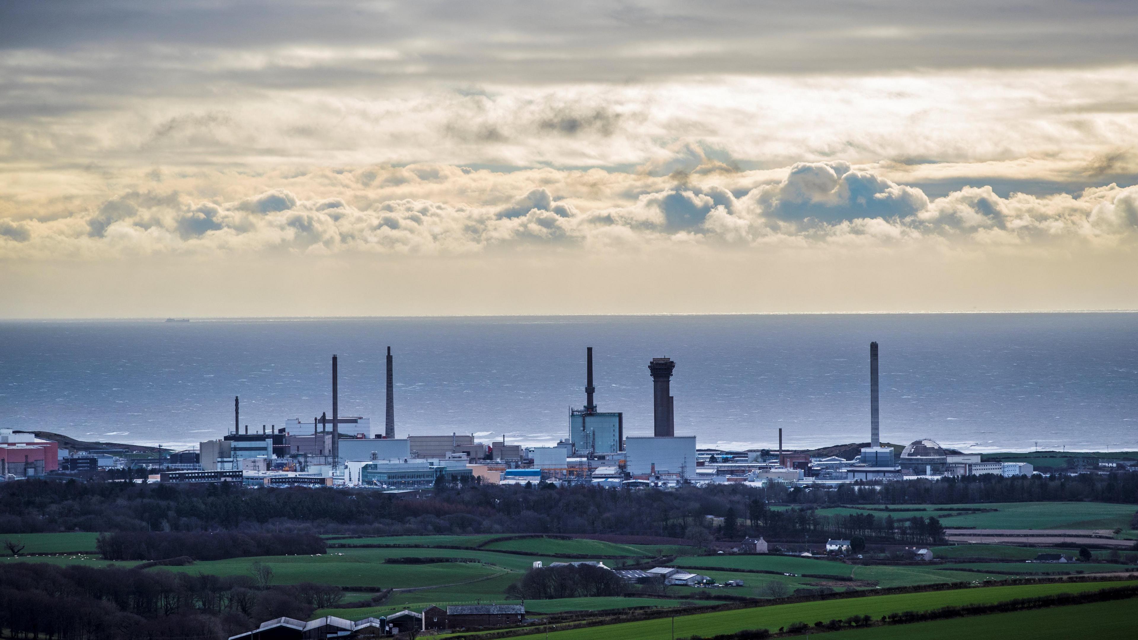 An aerial image of the Sellafield site near Seascale. There are green fields and open countryside in the foreground, with the industrial buildings and chimneys of Sellafield by the shore and the sea in the background. 