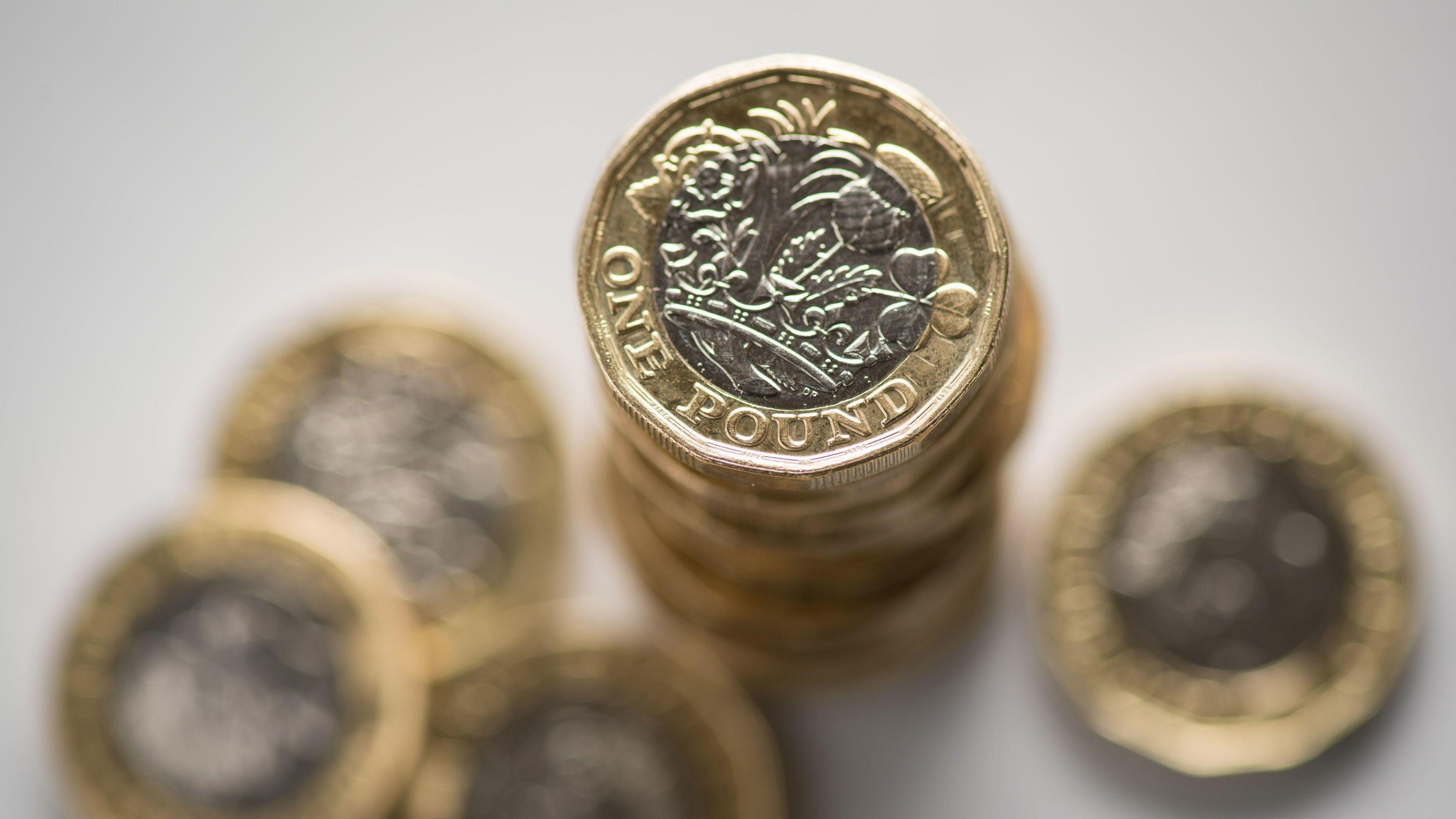 Close up image of a selection of British pound coins. The coins are stacked on top of one another. A couple of £1 coins are scattered around the stack. 