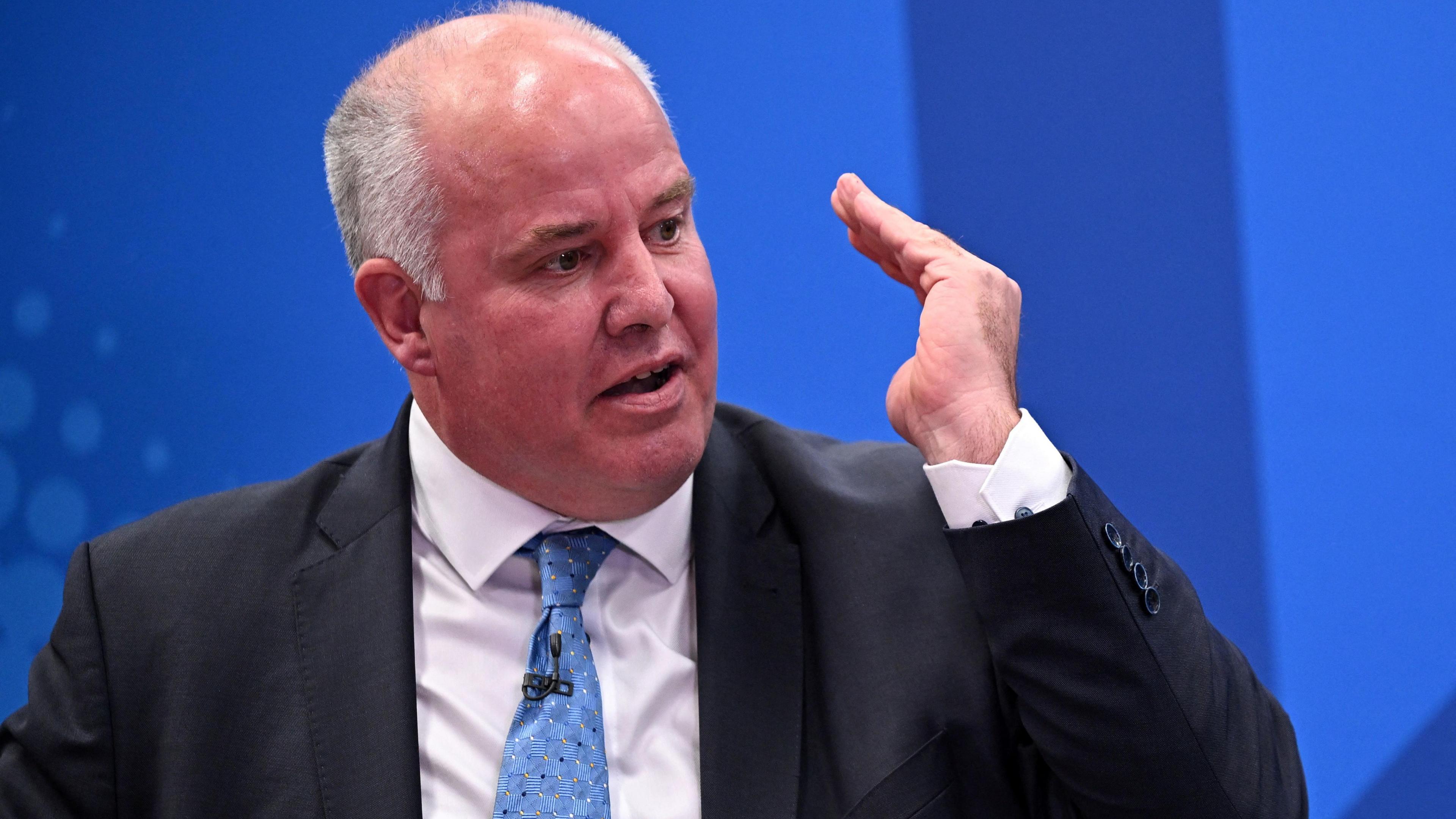 Andrew RT Davies sat in front of a blue background, with his hand gesticulating in the air. He is wearing a black suit, a white shirt and a blue tie.