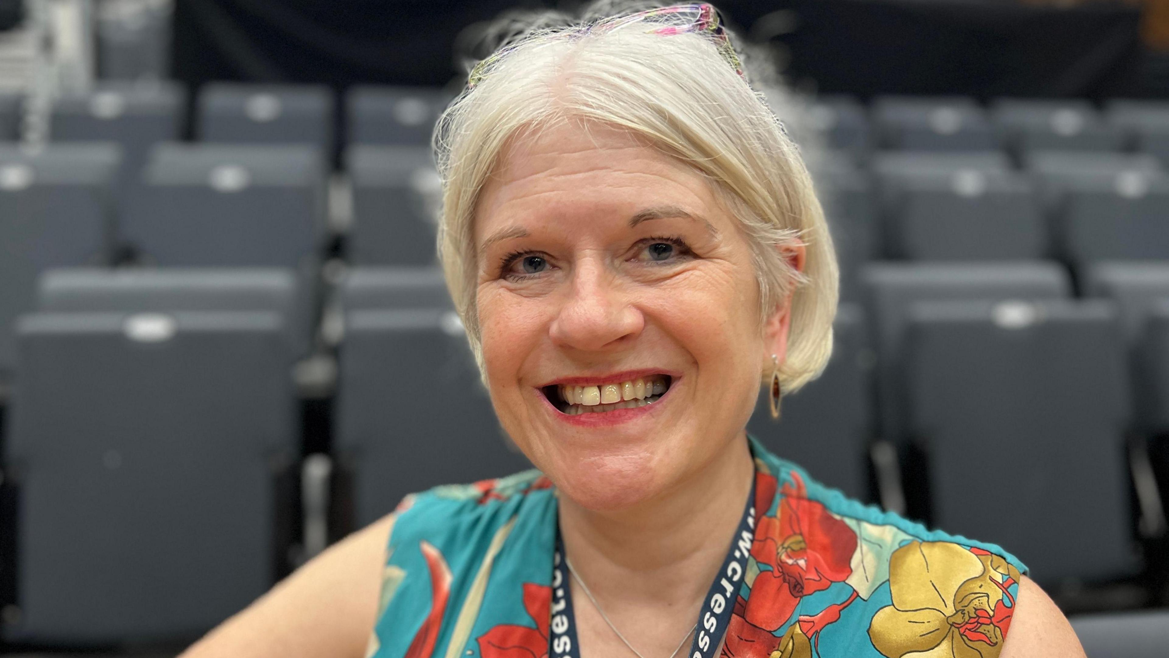 Ms Chantal is sat in the theatre seating smiling at the camera. She has medium length blonde hair, blue eyes and pink lipstick. She is wearing earrings and blye, red and gold flower print dress. She is also wearing glasses on top of her head.