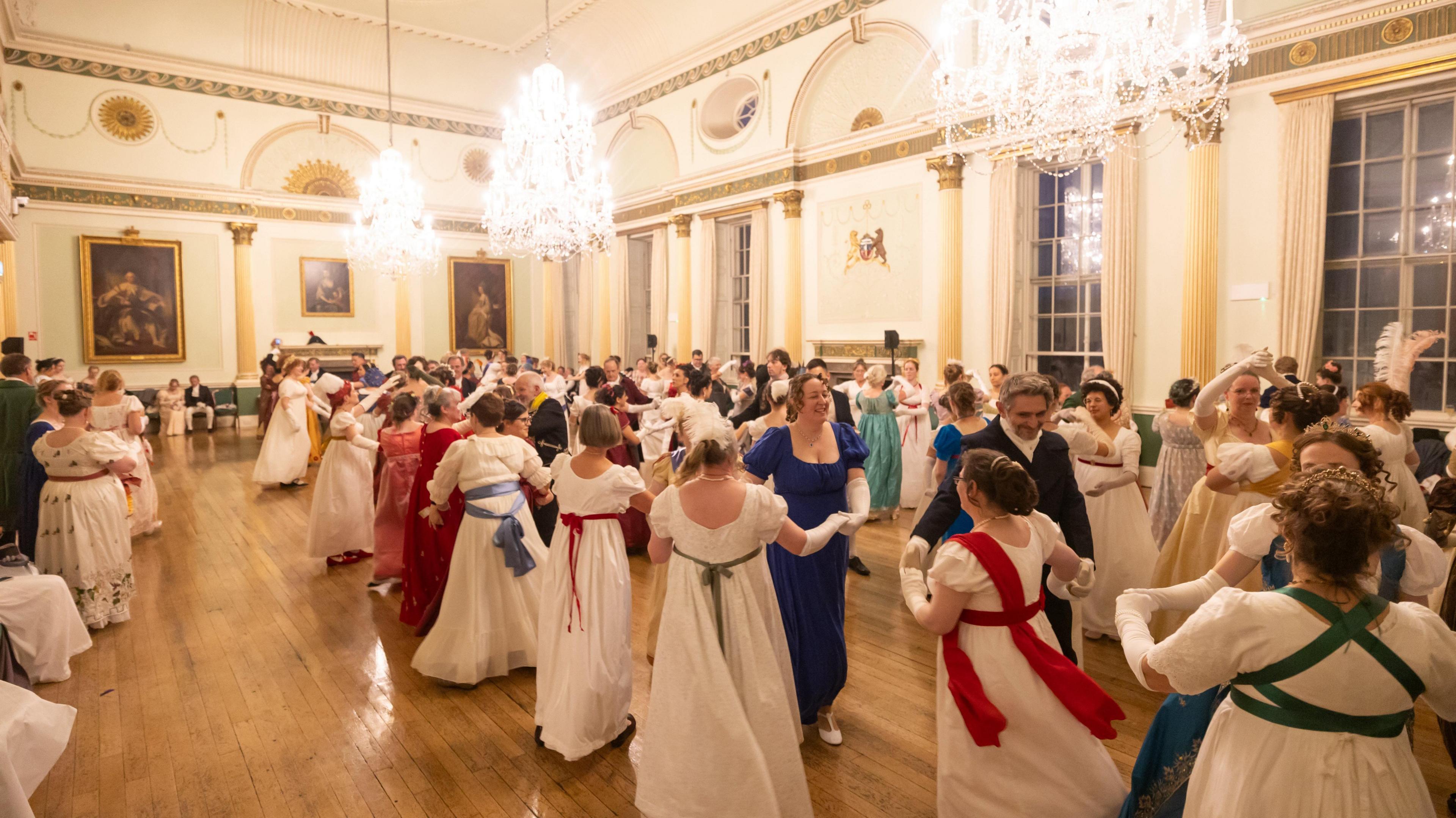 A ballroom full of people dancing while wearing regency costumes. They are all in couples. There are chandeliers hanging from the ceiling.