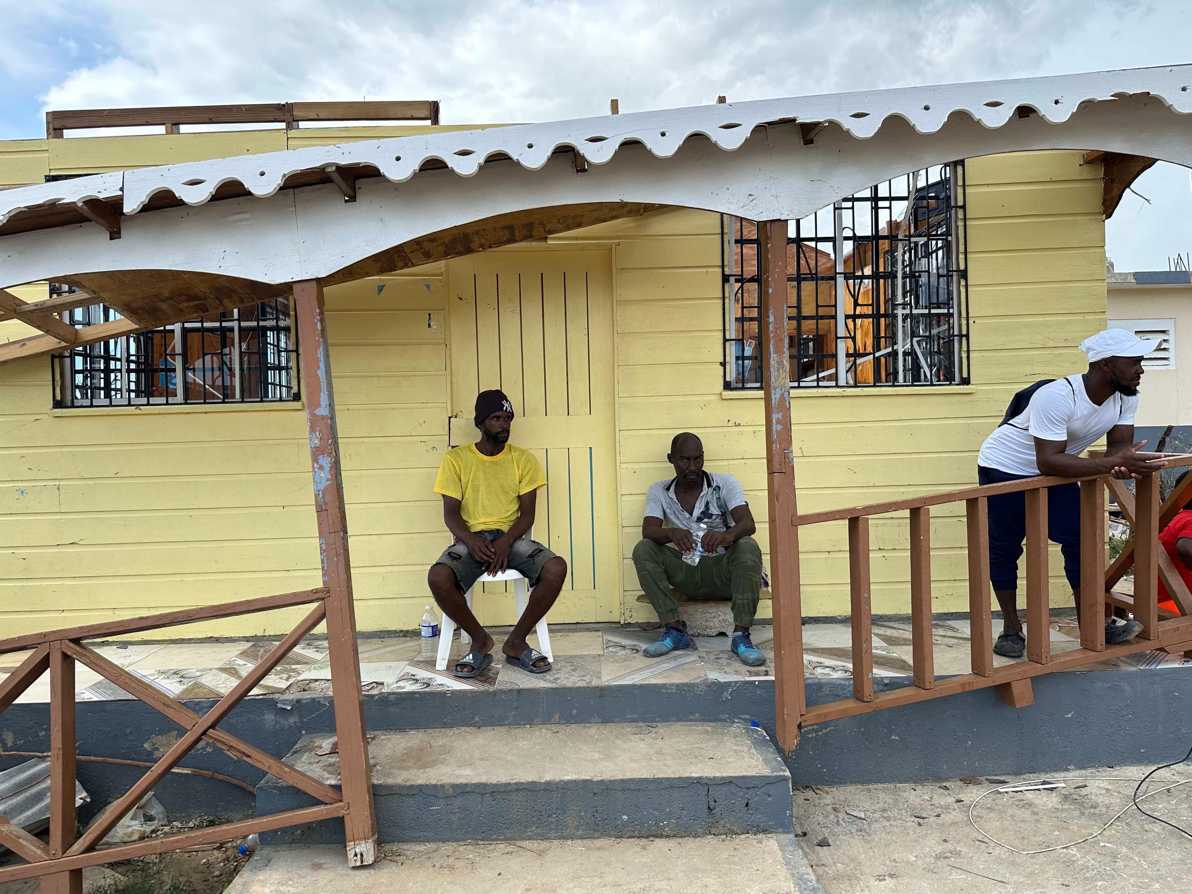 Two men are seated beneath a building half blown away by the storm