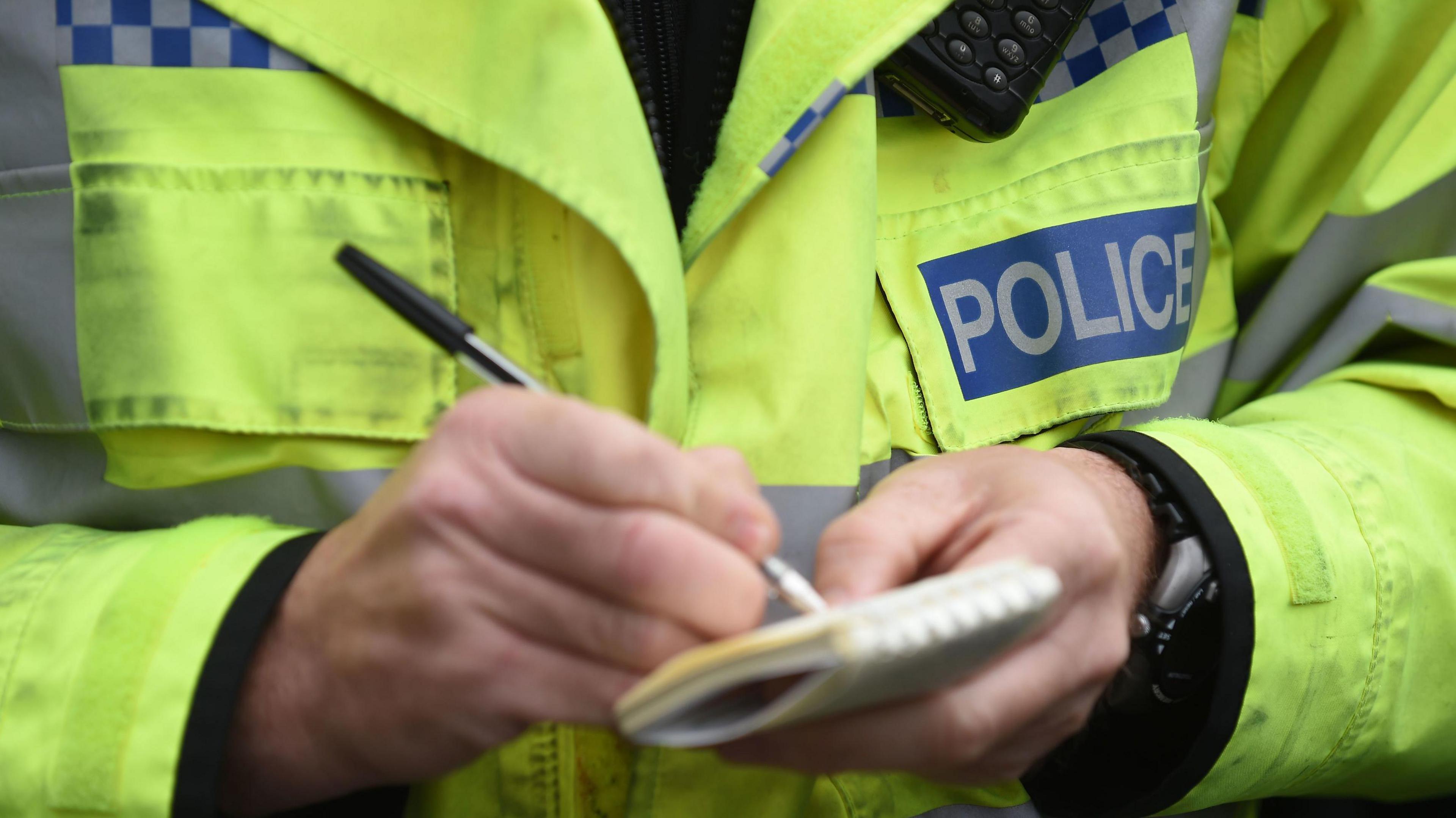 A close-up of a police officer wearing a bright yellow high-vis jacket with 'police' label on the front right breast pocket and above this is a phone clipped on underneath the collar of the jacket. He is holding a pen in the left hand and writing in a notebook, which he is holding with his other hand