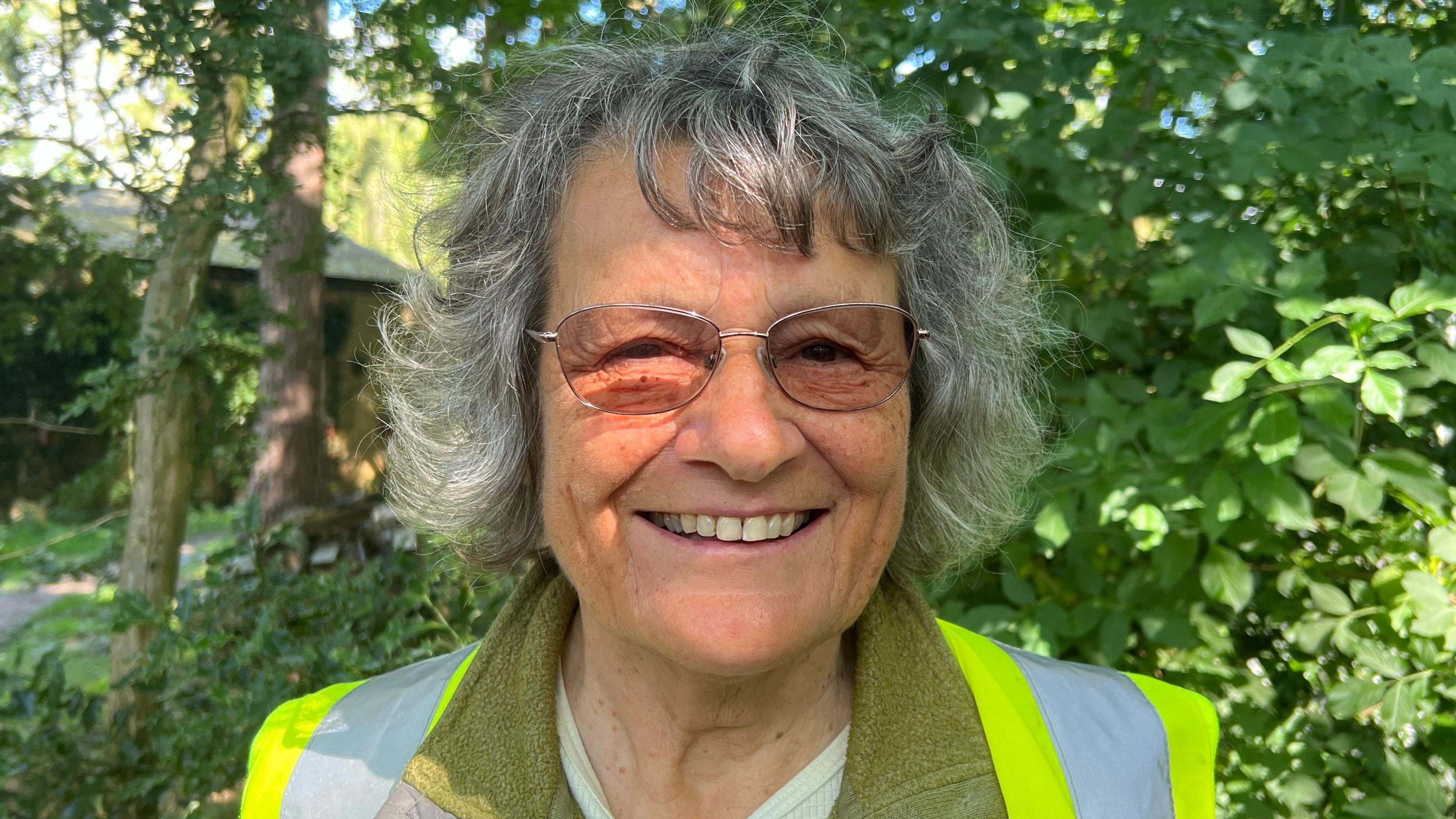 Volunteer Gill Reid, grey-haired and wearing spectacles and a hi-vis jacket, against a backdrop of trees 