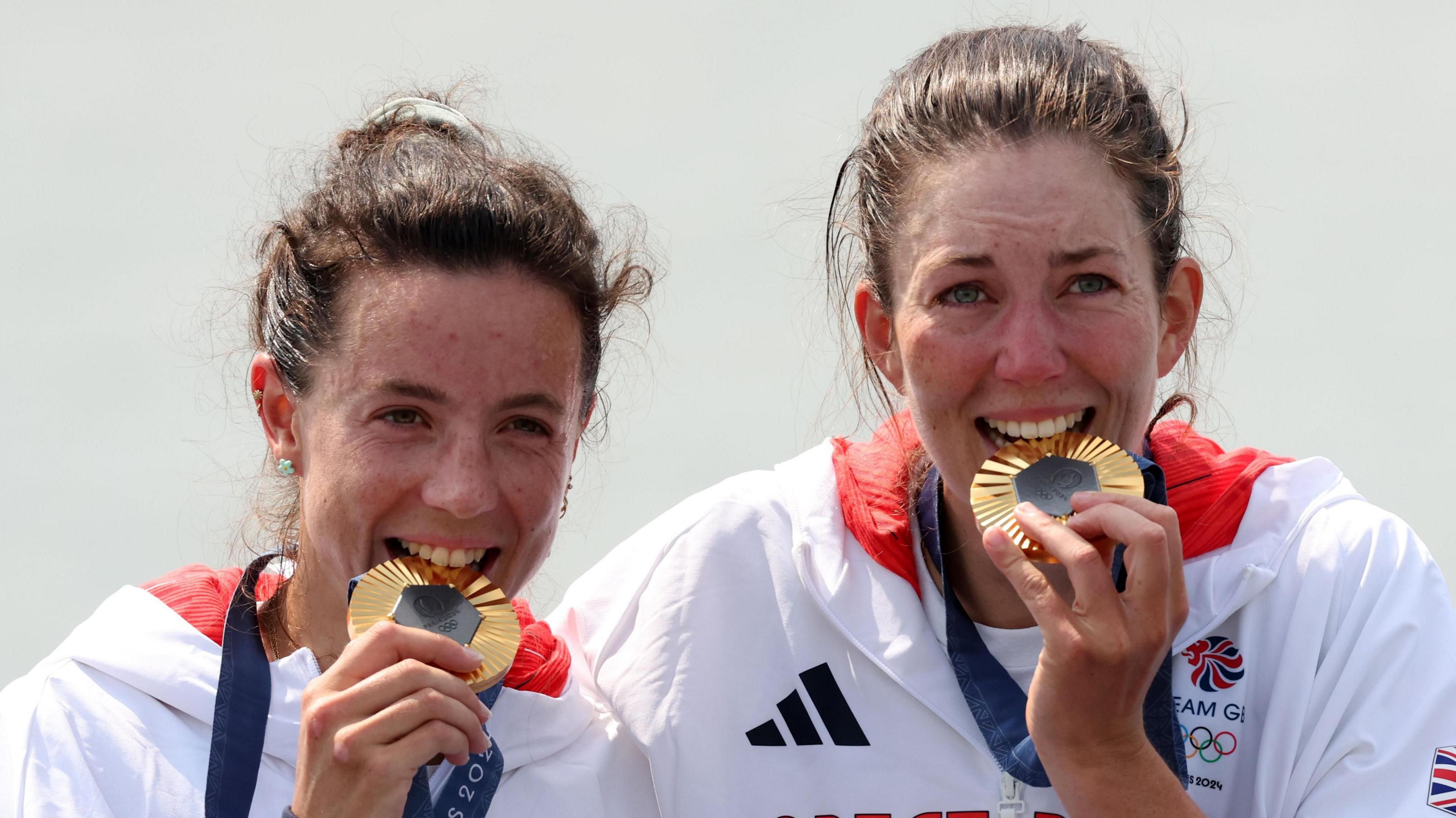 Two women in Team GB white jackets biting on gold Paris Olympics medals