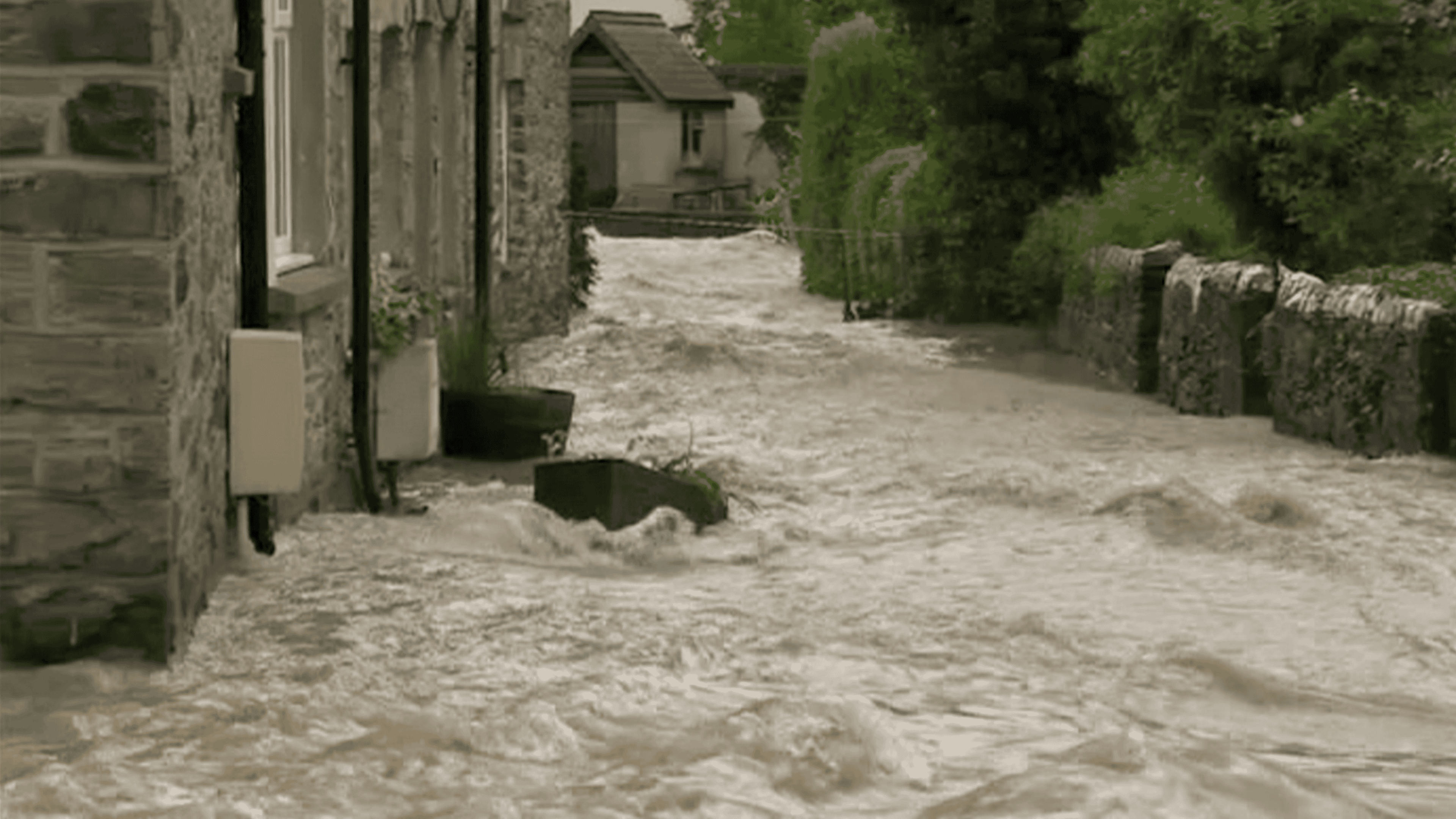 Llun o stryd yn Nhal-y-bont gyda dwr dwfn y llifogydd yn llifo trwyddi - mae un potyn blodau pren yn arnofio ar wyneb y dwr 