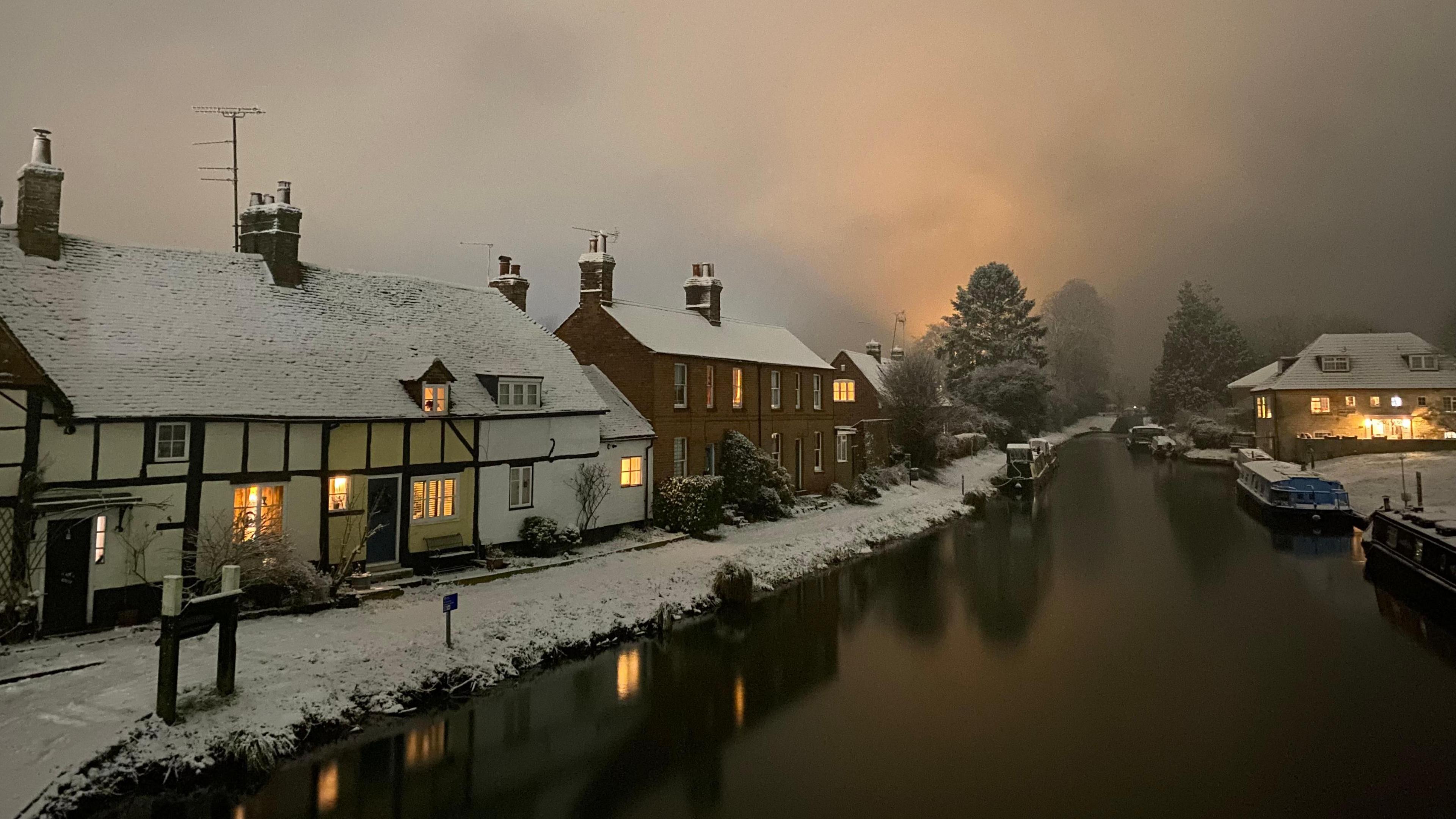 A row of snow-topped houses along a river, with boats moored up on the other side.