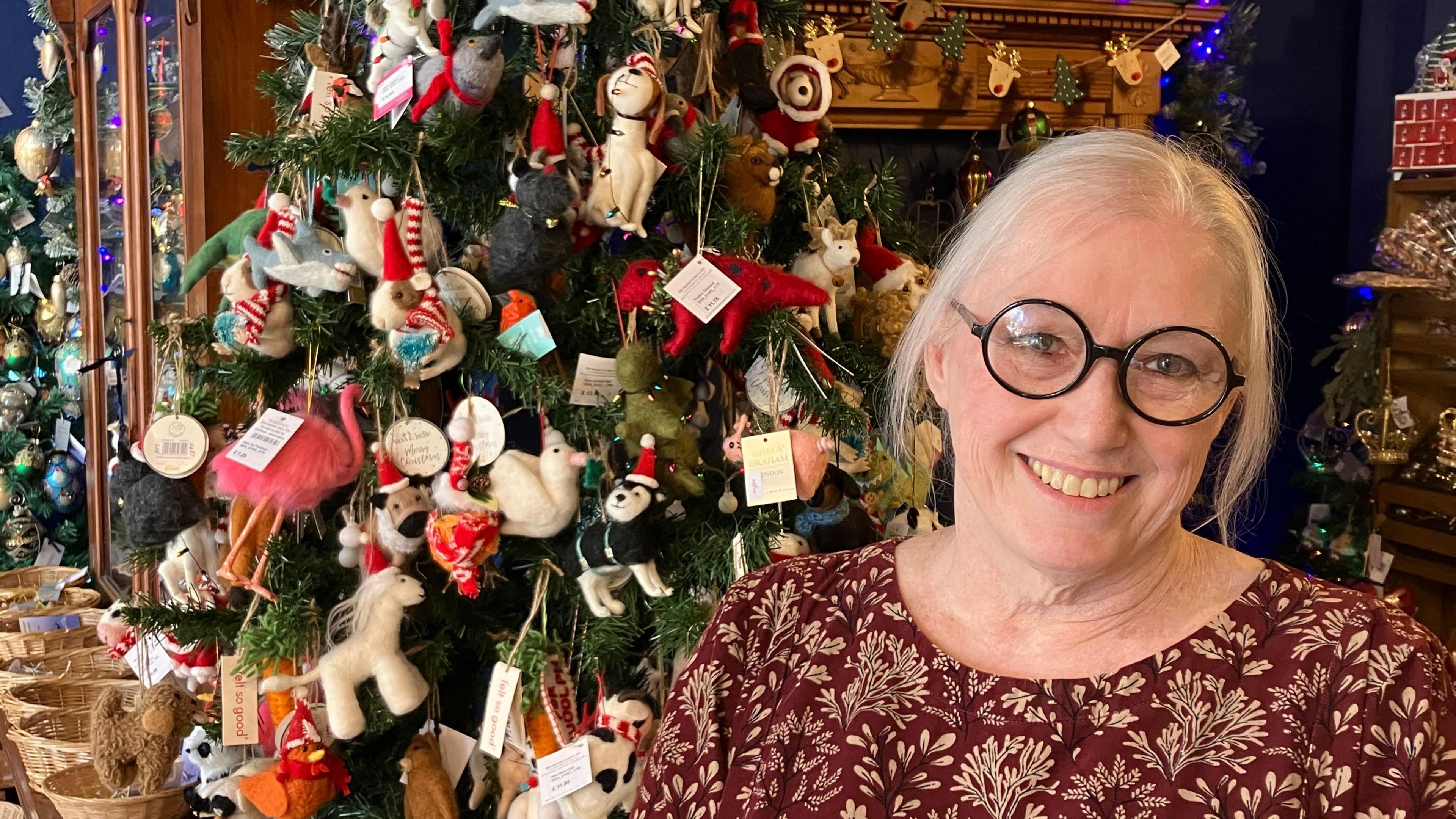 A person wearing glasses and a red top stands in front of a Christmas tree covered with bright decorations 