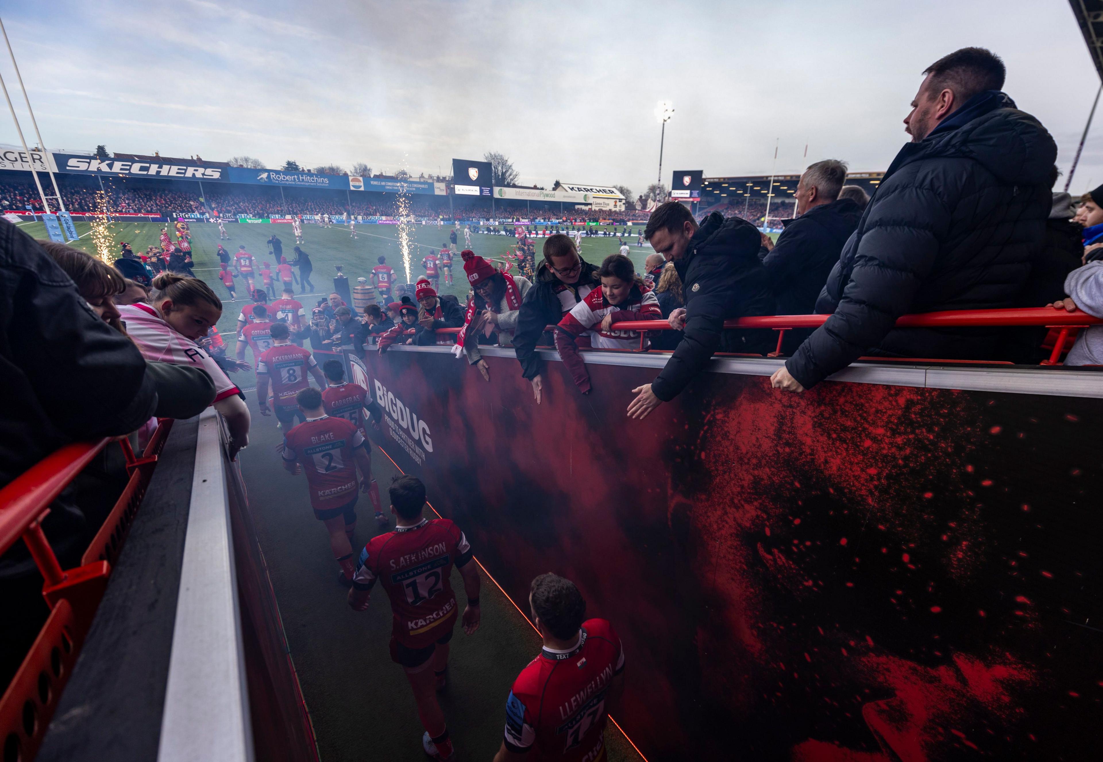 Fans lean down from the stands to try to high-five Gloucester Rugby players as they walk out of the tunnel and onto the pitch. Pyrotechnics can be seen going off on the field in the background.