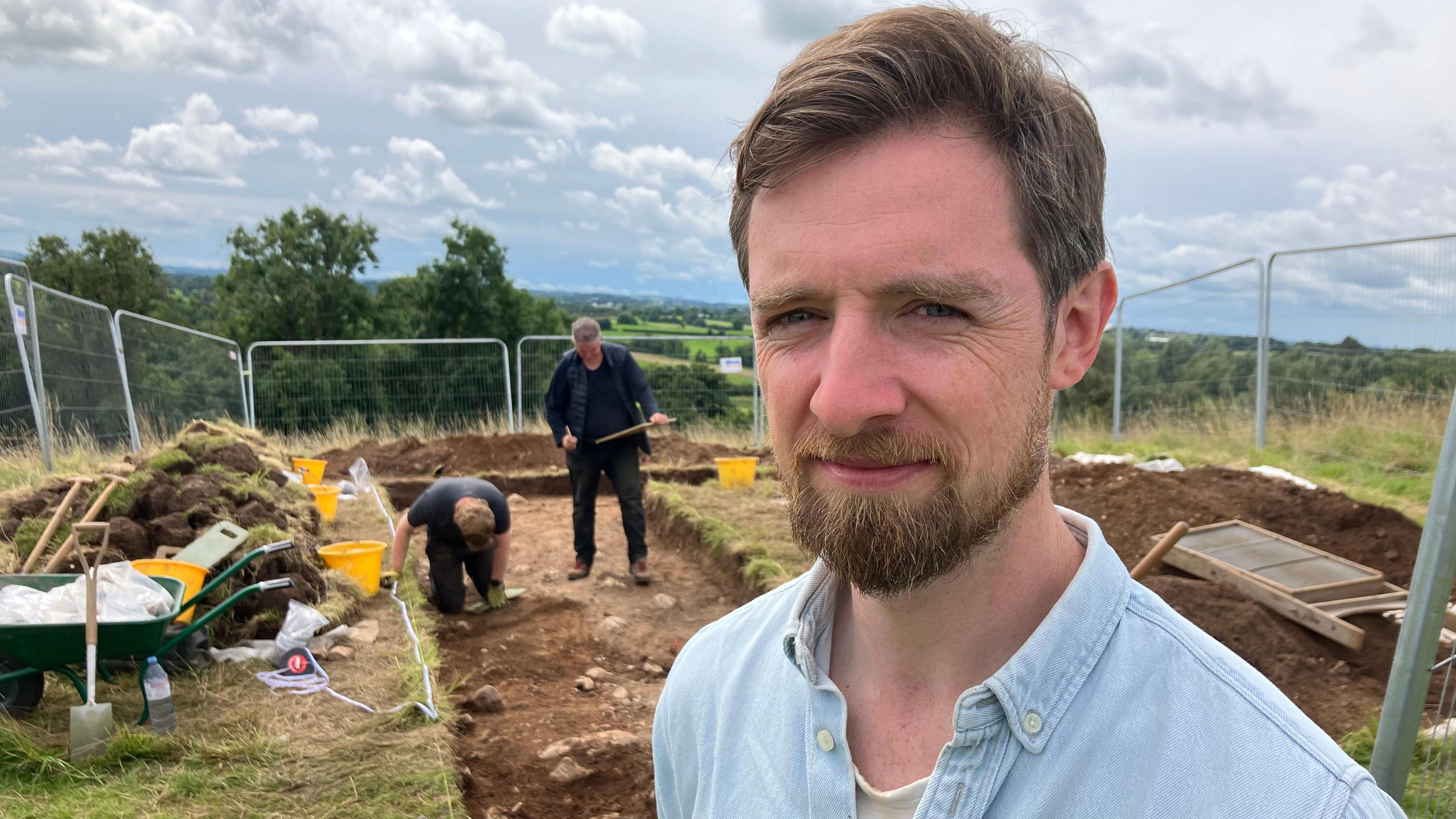 Dr Patrick Gleeson is standing in front of two archaeologists who are working on soil in a field. Grass has been pulled off. A shovel and a wheelbarrow are next to a pile of grass. Dr Gleeson is wearing a denim top.