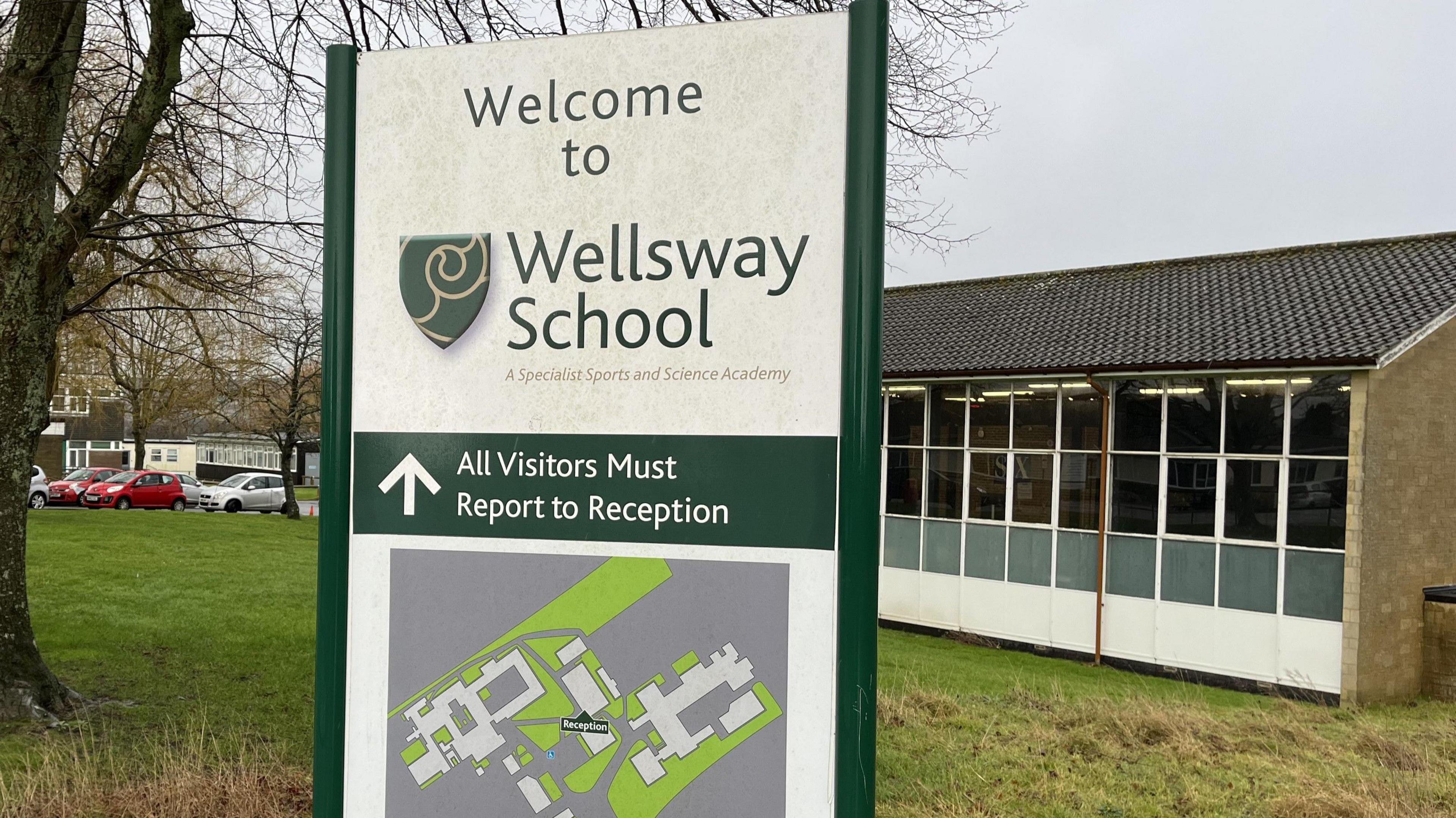 A welcome sign outside Wellsway School, directing visitors towards reception. In the background there is a small car park, a grassy section with trees, and a building with a tiled roof and lots of windows.