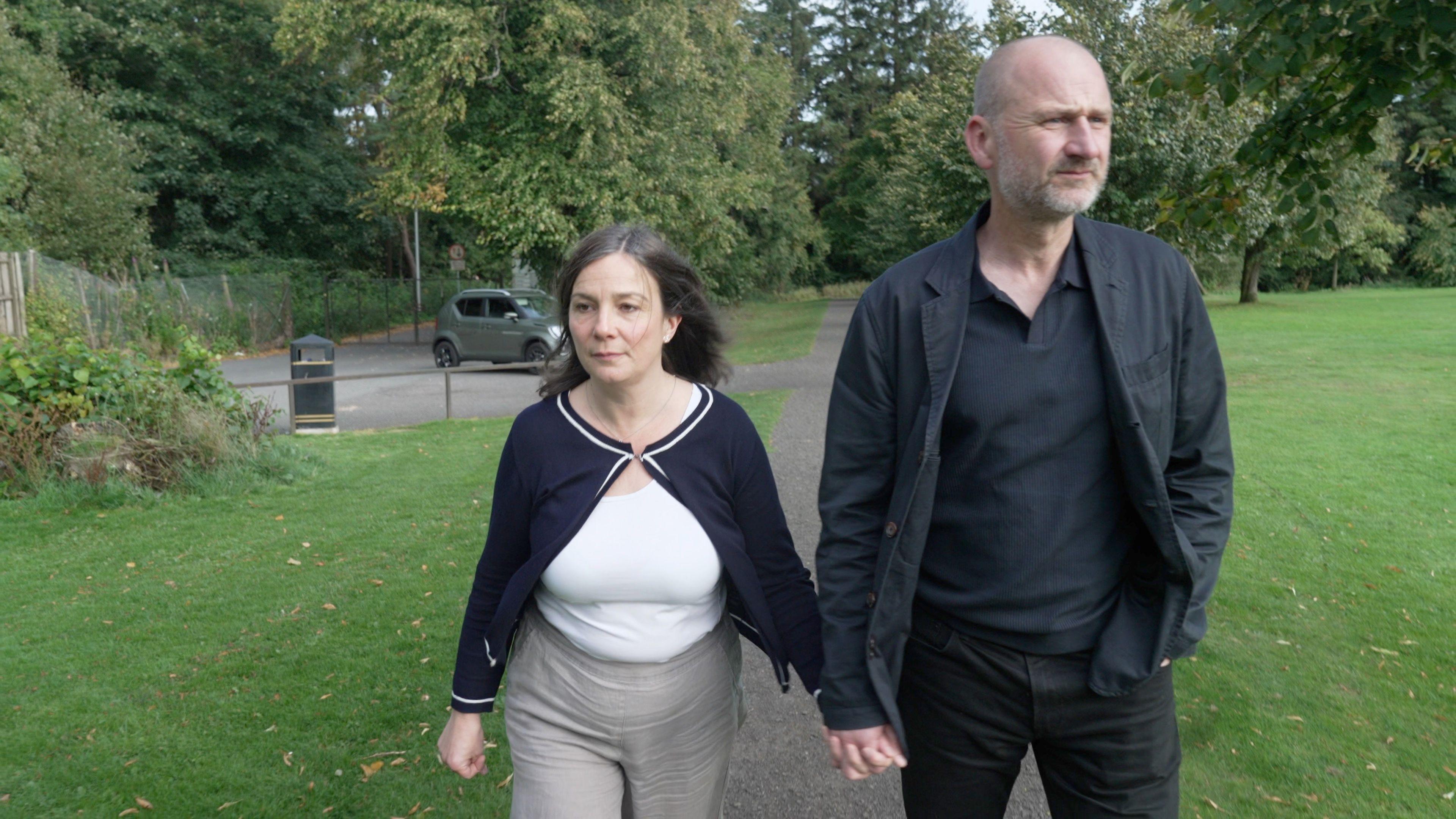 Ros and Mark holding hands as they walk in a park. Mark is wearing a black shirt and jacket. Ros has a white top on with a black jacket.