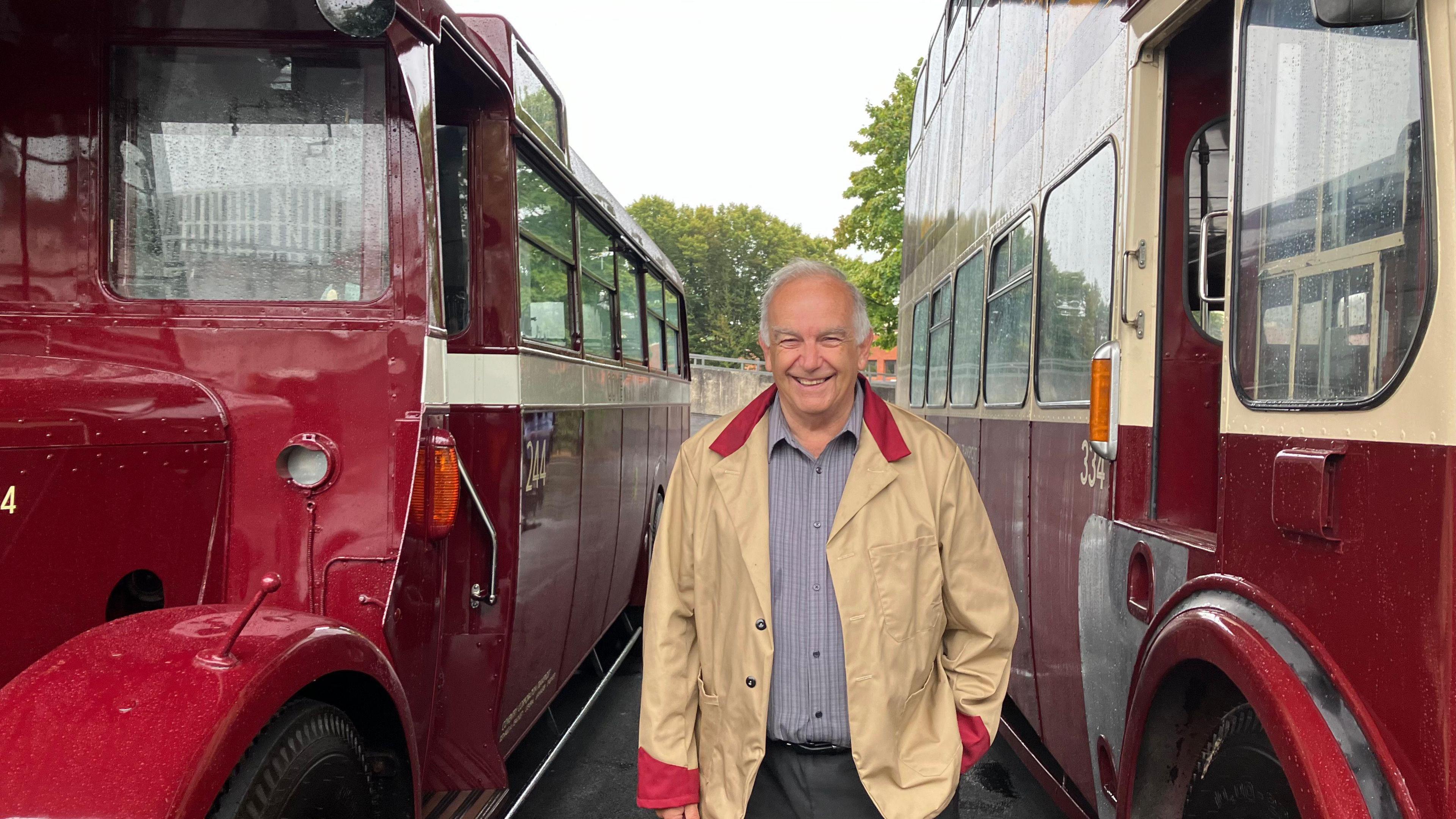 A man in a light brown coat with a red collar and cuffs, smiles as he stands between two historic red buses