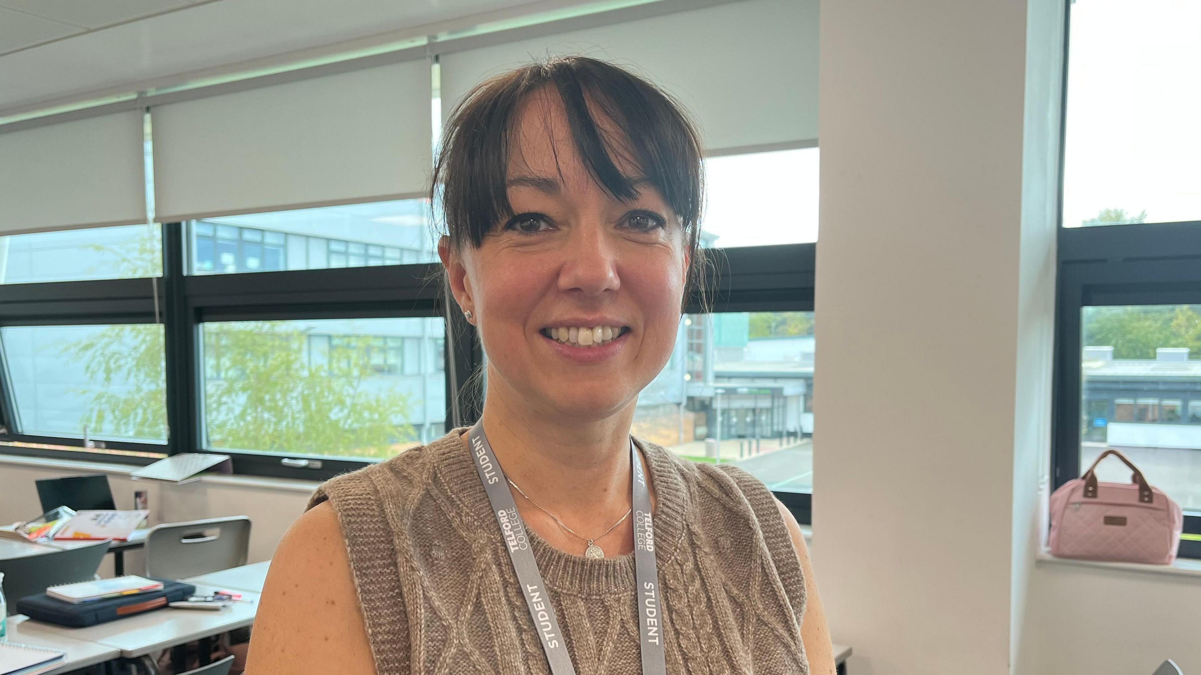 Emma looking into the camera and smiling. She's wearing a light brown sleeveless jumper, and has a grey Telford College lanyard. The room has white walls and grey chairs and desks. Out of the window you can see some more modern buildings on the campus. The desks are covered in pencil cases, folders, and water bottles.