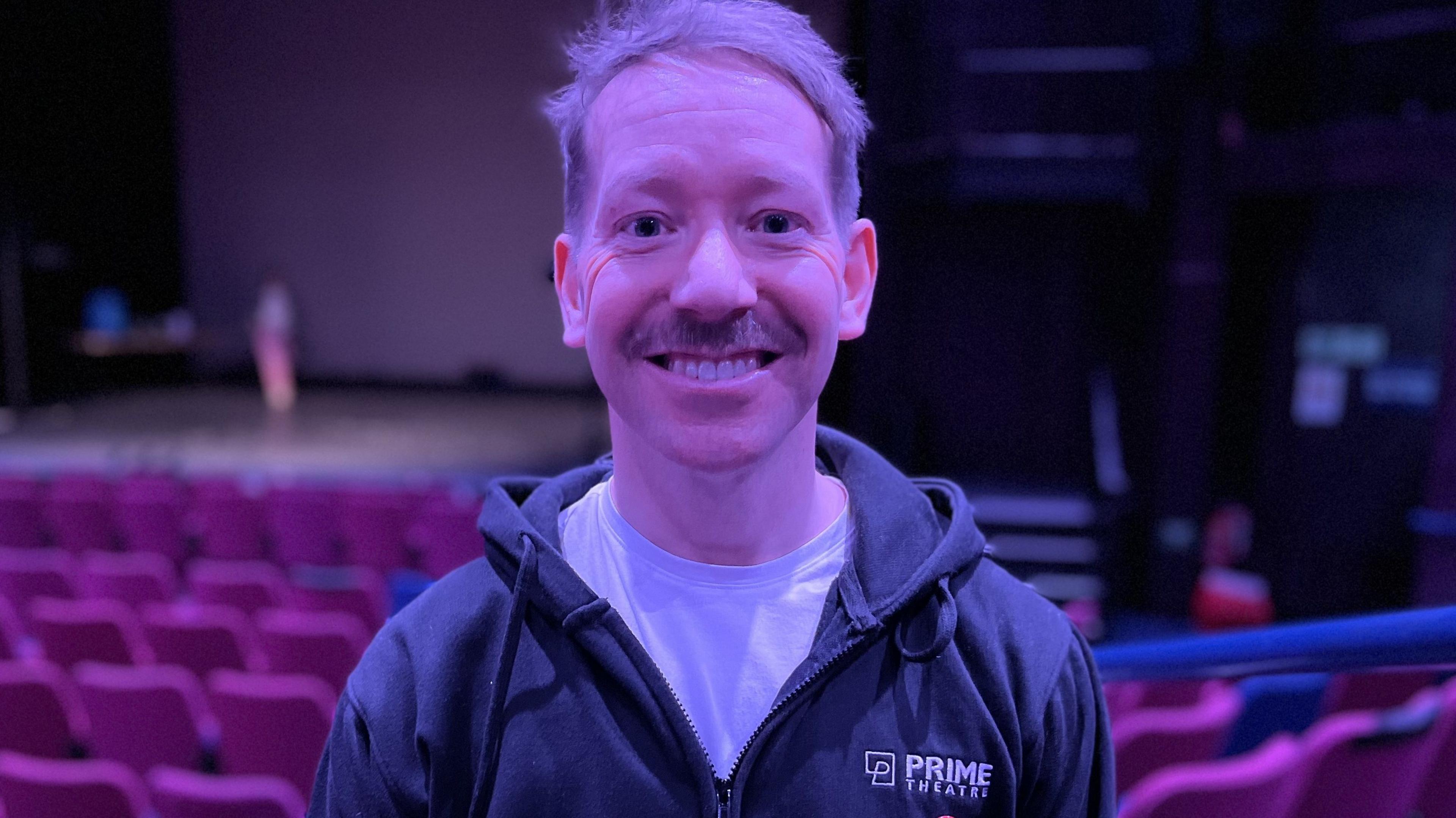 Mark Powell, Artistic Director at Prime Theatre smiling at the camera wearing a navy blue Prime Theatre hoody