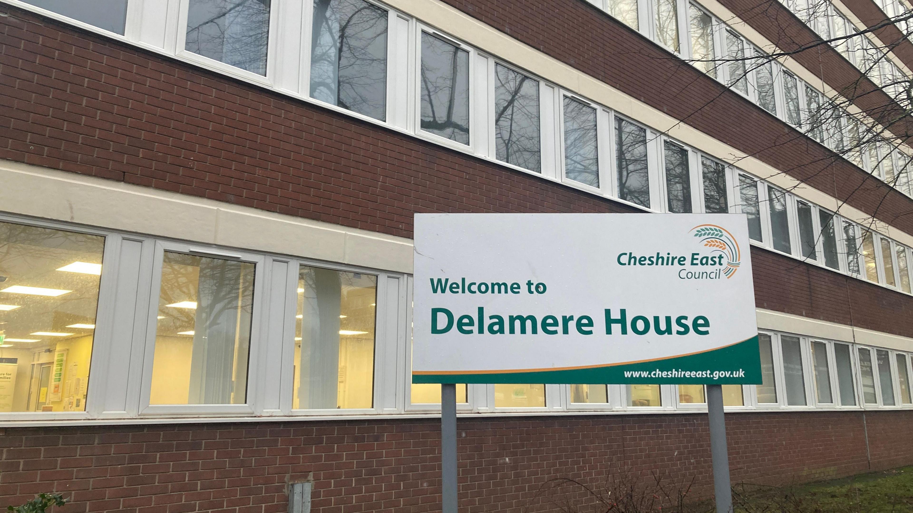 An angled shot of Delamere House in Crewe, Cheshire East Council's headquarters, with it's brick facade interlaced with long sections of white windows. The Delamere House green and white sign is pictured on the lawn in front of the building. 