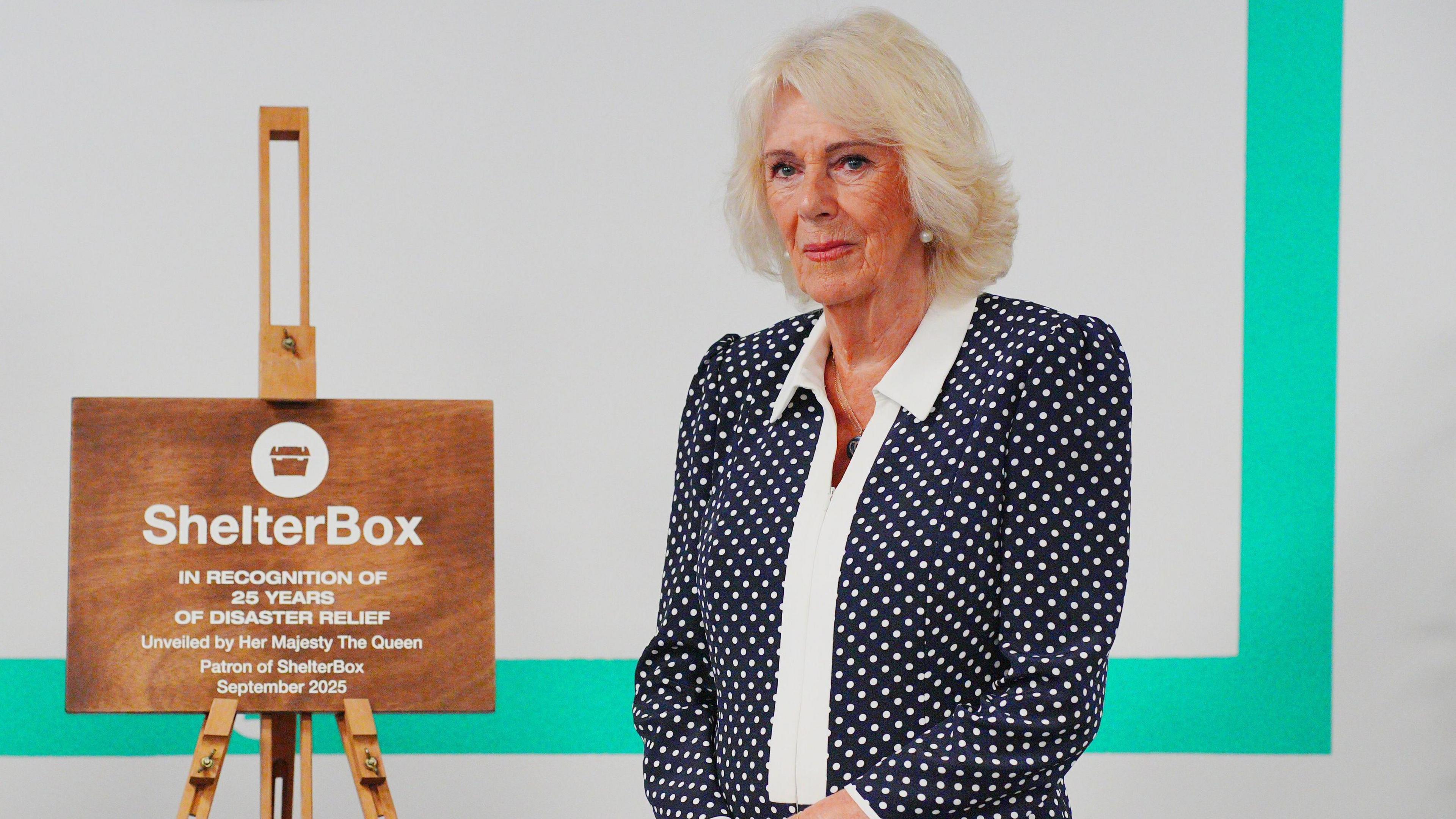Dressed in a dark dress with white polka dots and a white collar the Queen stands besides a wooden plaque which is standing on an easel. It reads ShelterBox In recognition of 25 years of disaster relief unveiled by Her Majesty the Queen, Patron of ShelterBox September 2025.