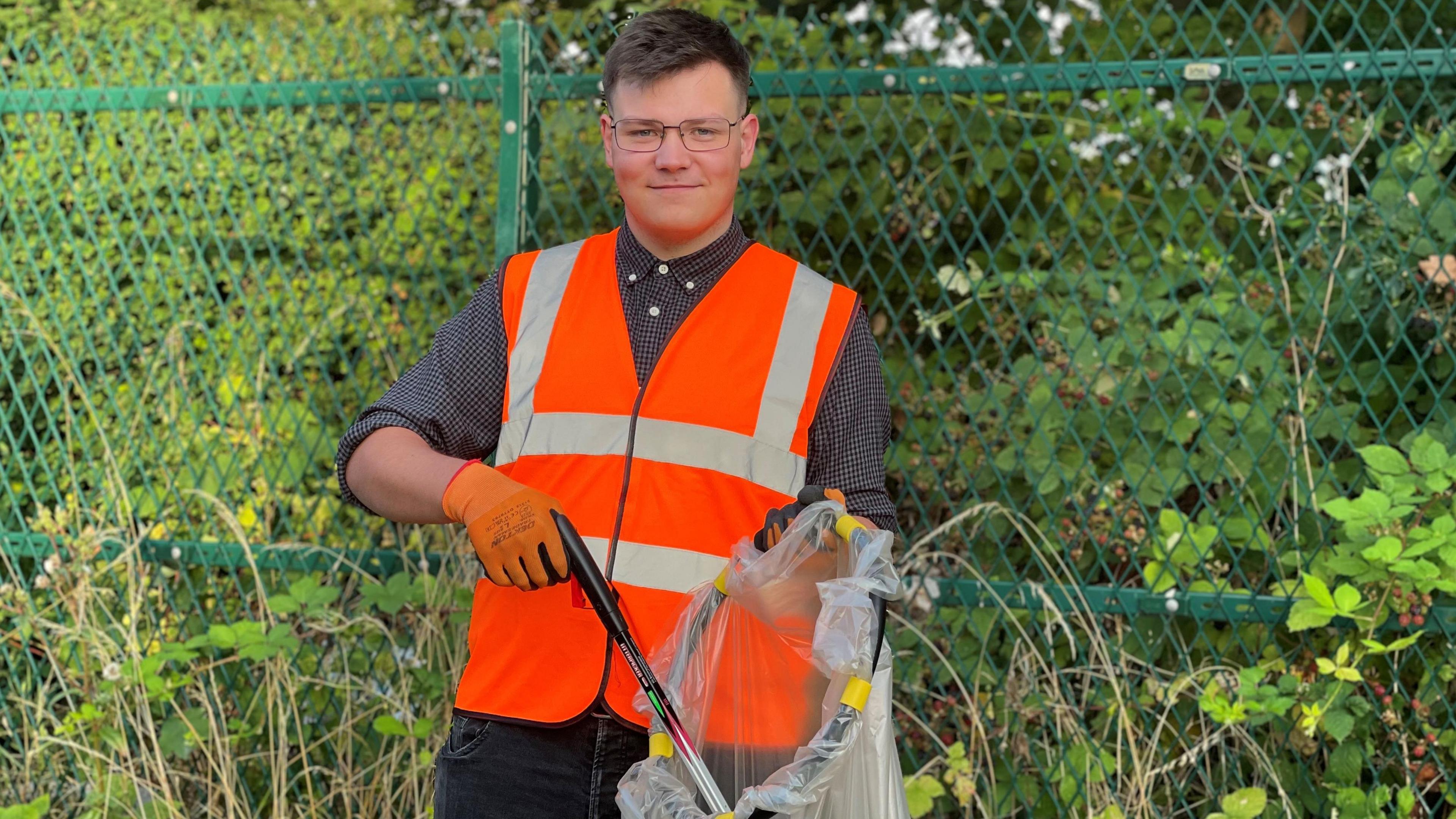 Man wearing glasses and a bright orange vest in front of bushes and woodland.
