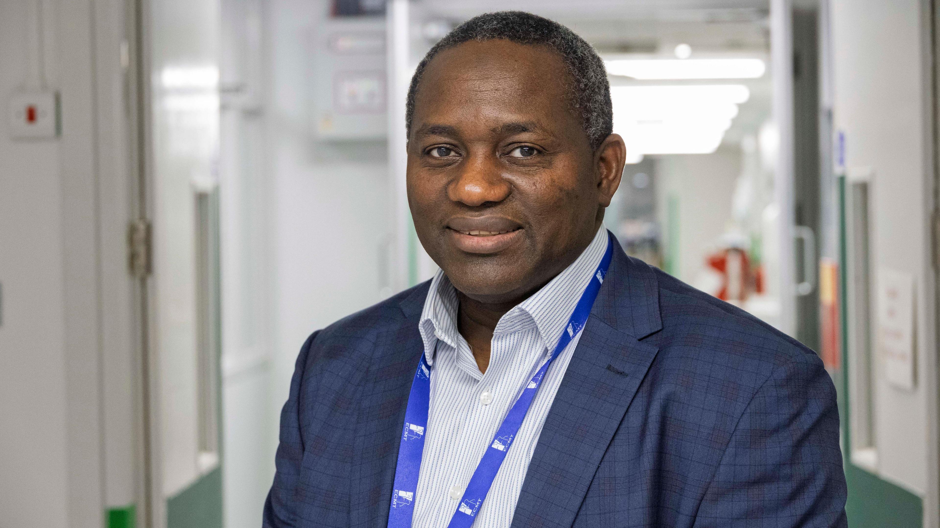 A portrait photo of a man posing for the camera with his hands held at his front, wearing a navy blazer and white shirt with a lanyard on. He is stood in a hospital corridor.