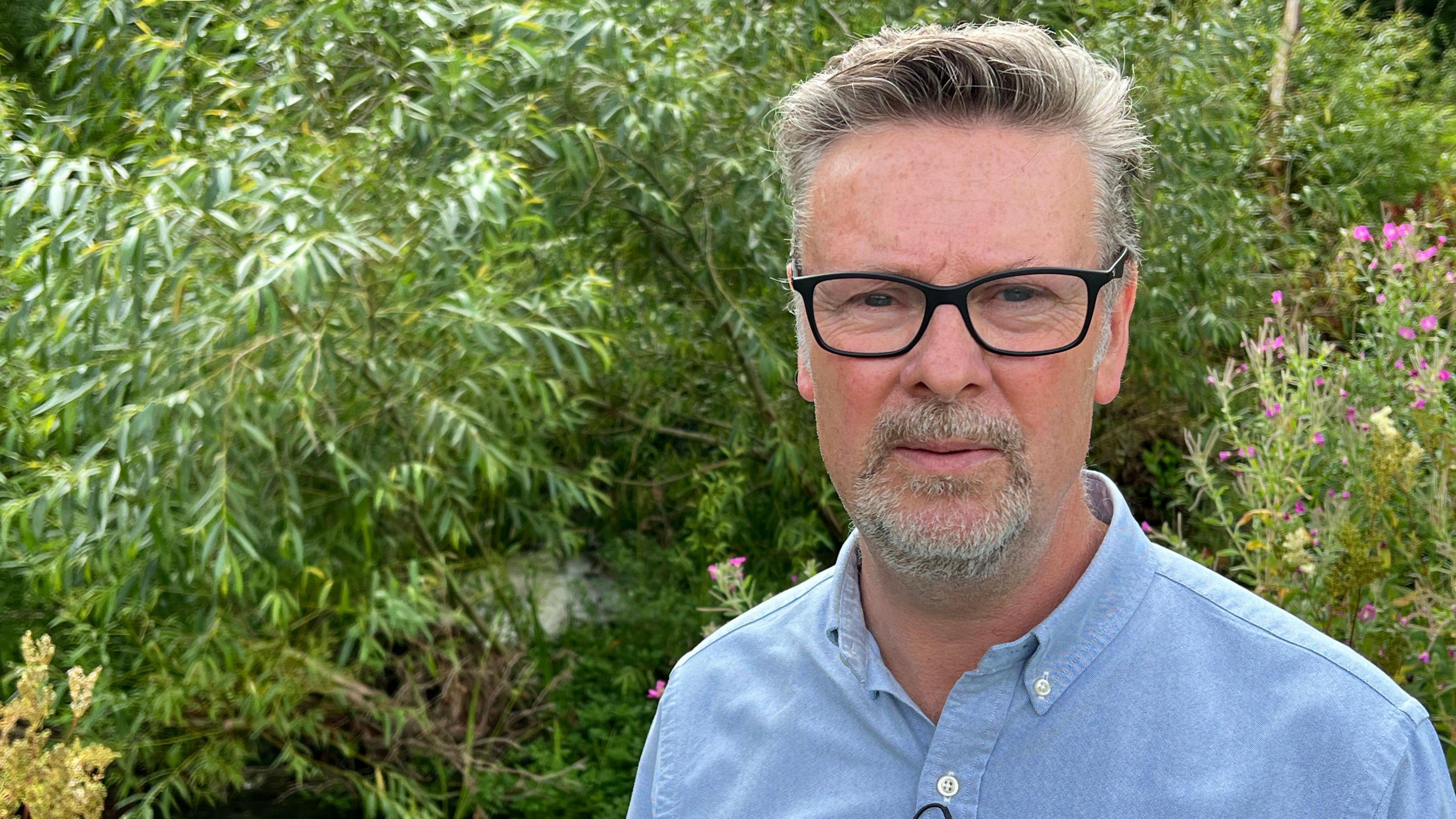 Councillor Tristan Wilkinson stands in front of green bushes and trees. He is wearing a blue shirt and black framed glasses, and has grey hair and a grey beard.