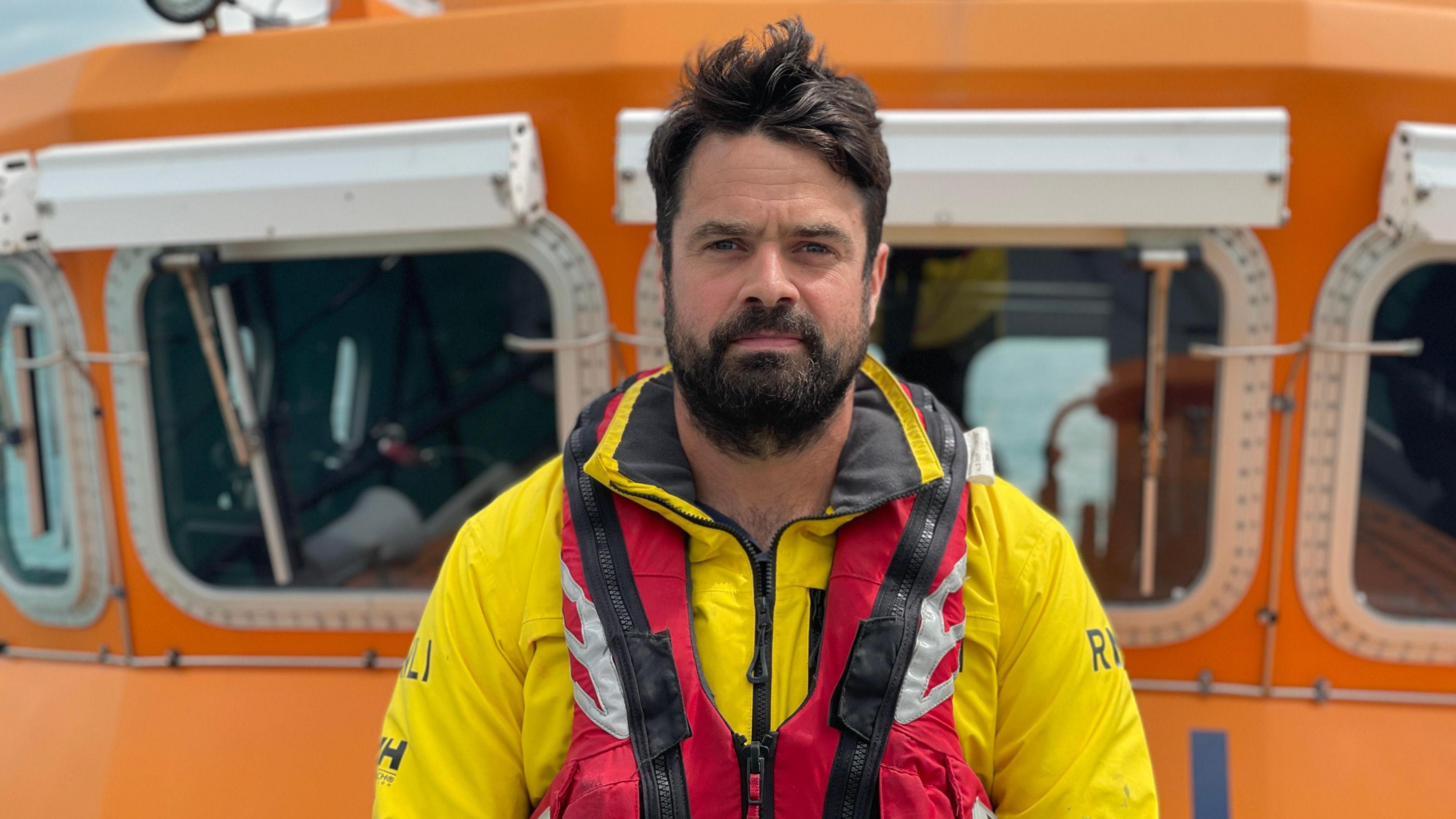 Dan Sinclair - a man with black hair, a black beard and a black moustache. He is wearing a yellow RNLI wetsuit and a red life jacket. The picture has been taken from a boat in the sea.