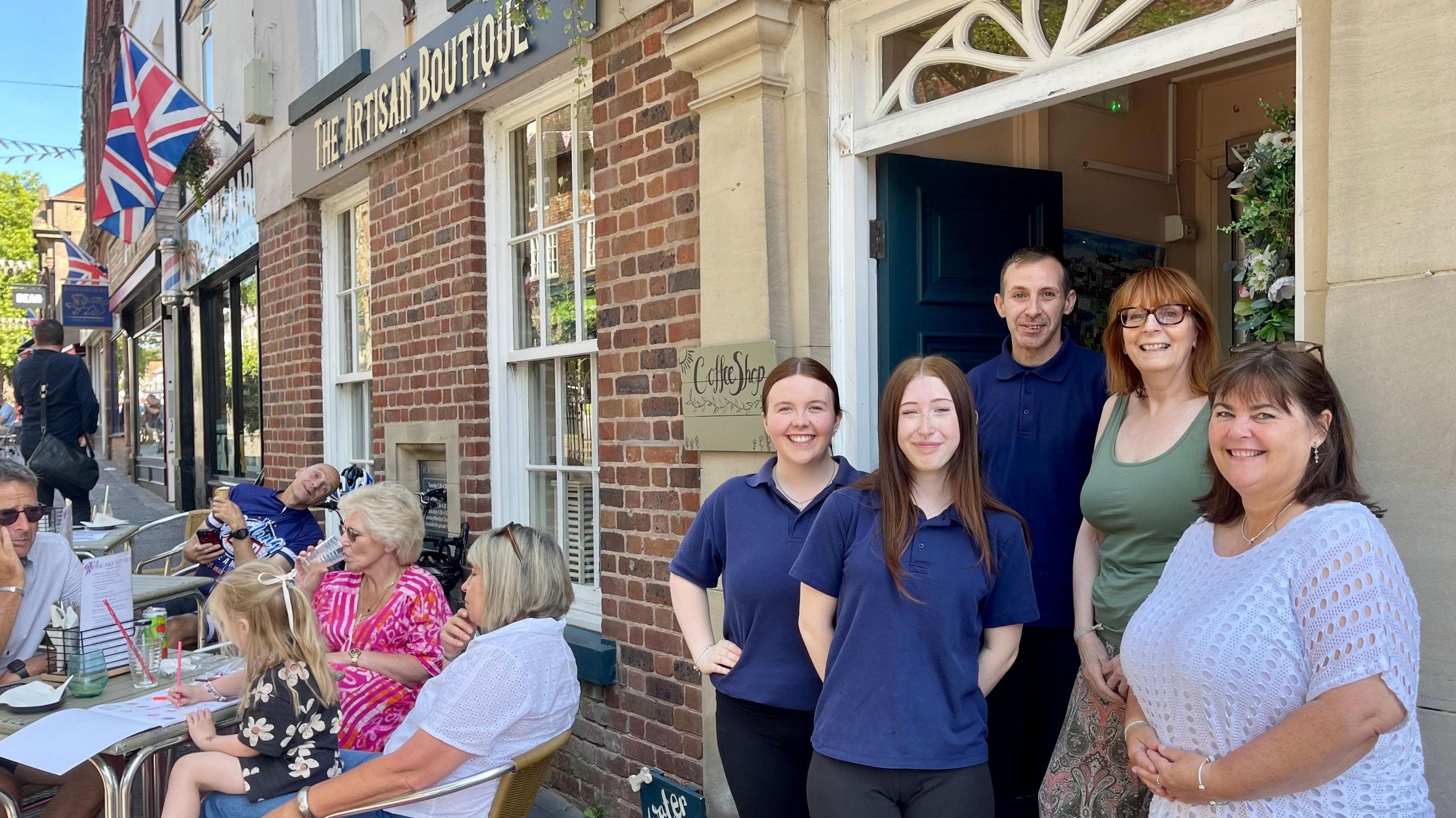 Four women and one man stand in the doorway of a cafe. Outside, people are sat at outdoor tables. The sign above the face reads The Artisan Boutique.