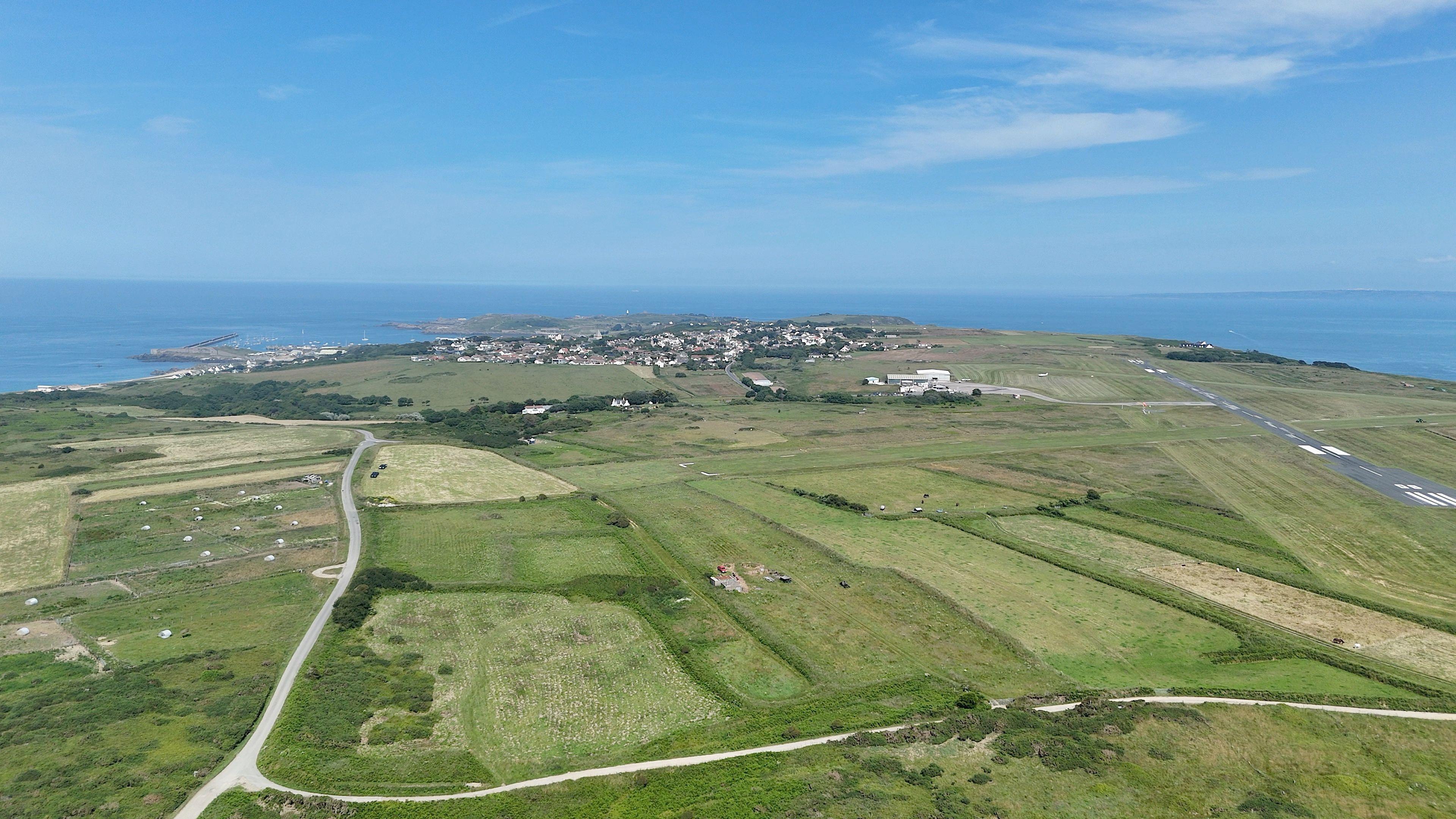 Aerial shot of green fields in Alderney. A large road runs through the fields and leads to a residential area in the distance. The sea surrounds the island.