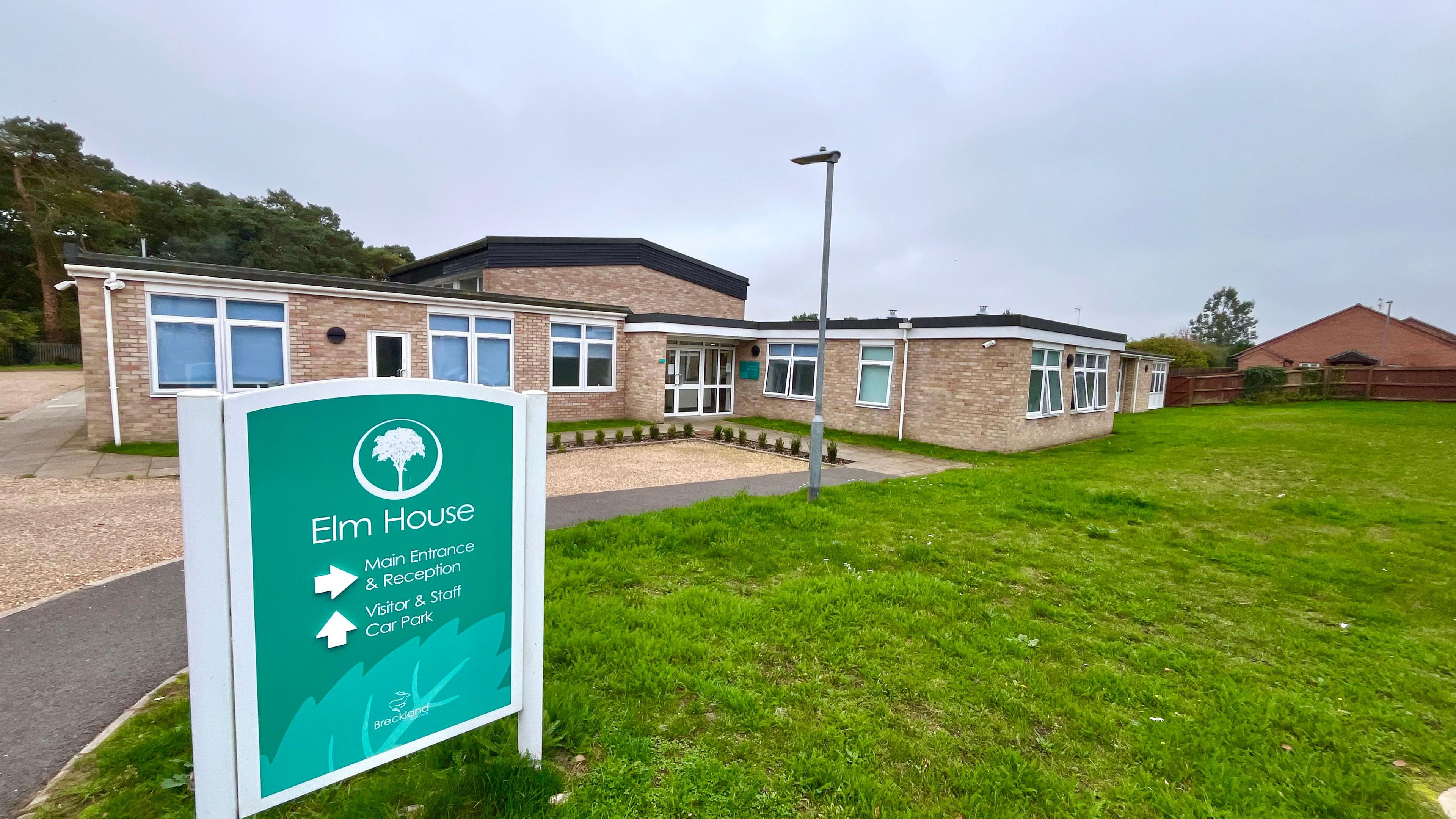 The outside of Elm House, which is a single-storey building with windows either side of a main entrance. There is a green sign at the front of the picture with its name and the logo of an elm tree on it