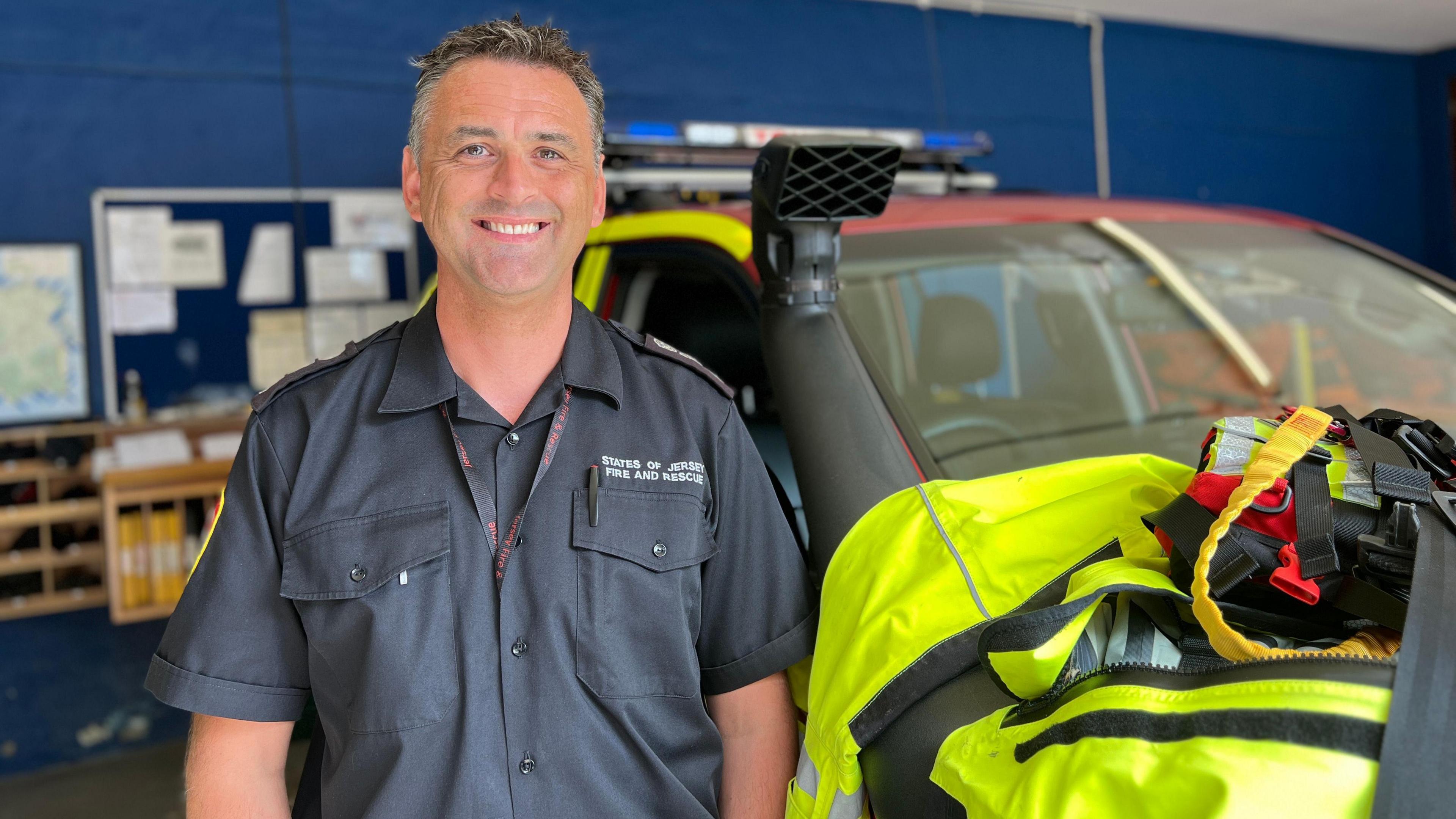 Ryan Hall smiles at the camera with his teeth showing. He is wearing a States of Jersey Fire and Rescue short-sleeved shirt with a lanyard tucked in between the buttons on his shirt. He is standing next to a rescue vehicle.