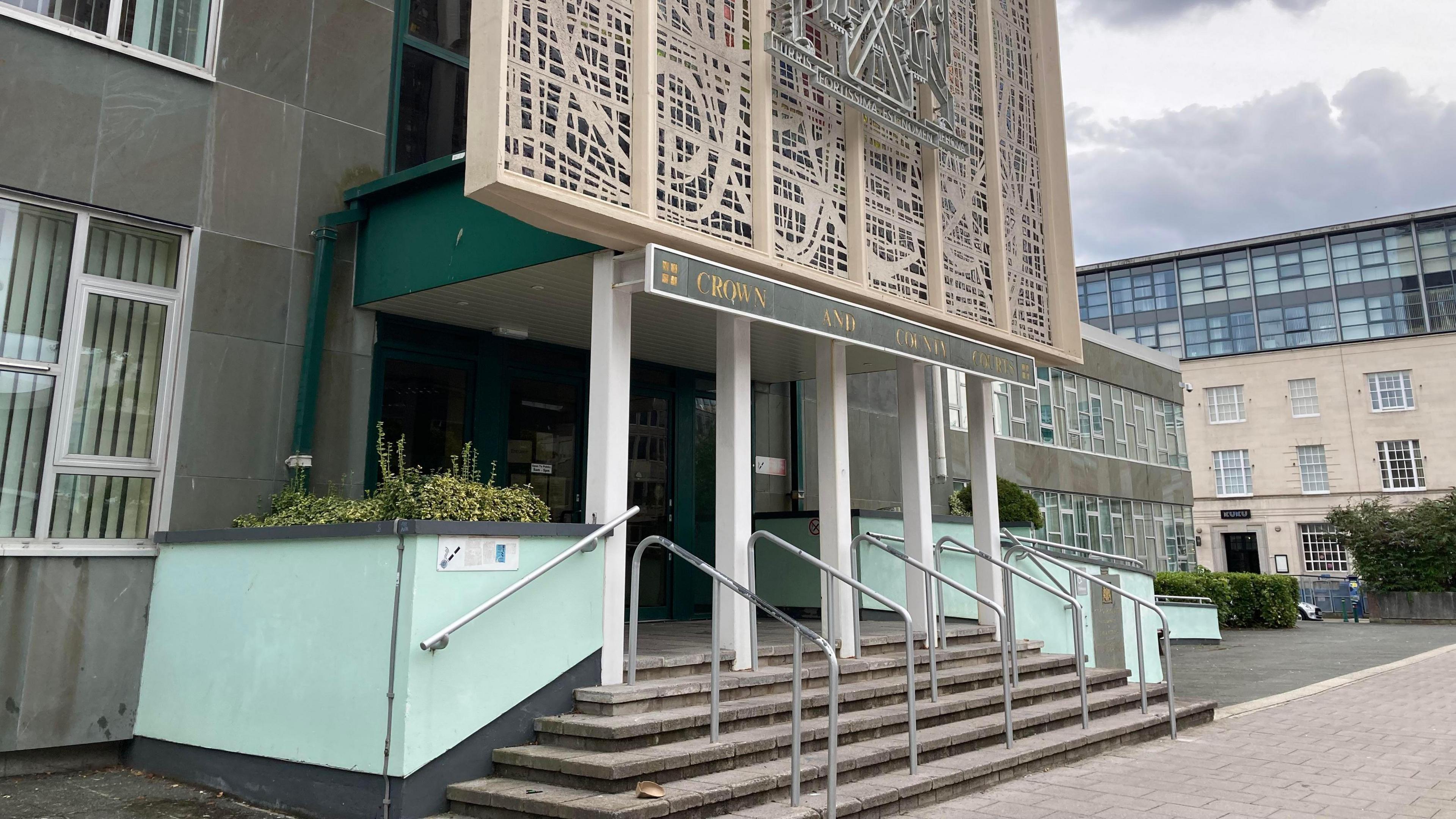 Entrance for Plymouth Crown Court. There are steps leading up to the main doors which have four thin, white pillars supporting the above floor.